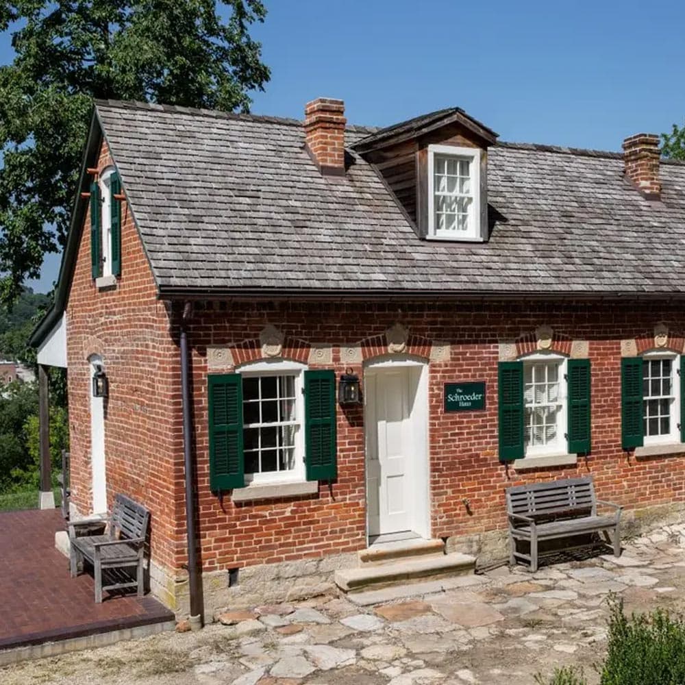 A brick house with green shutters and a sign reading "Schroeder House" sits under a clear blue sky.