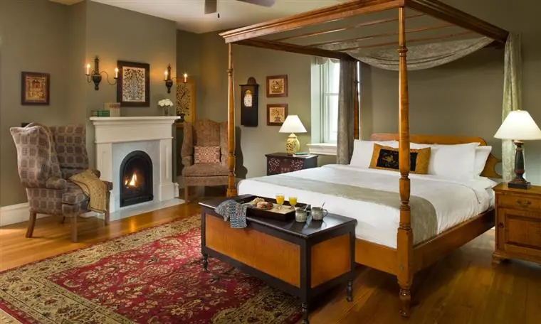 A cozy bedroom featuring a four-poster bed, armchairs, a fireplace, and a decorative rug.