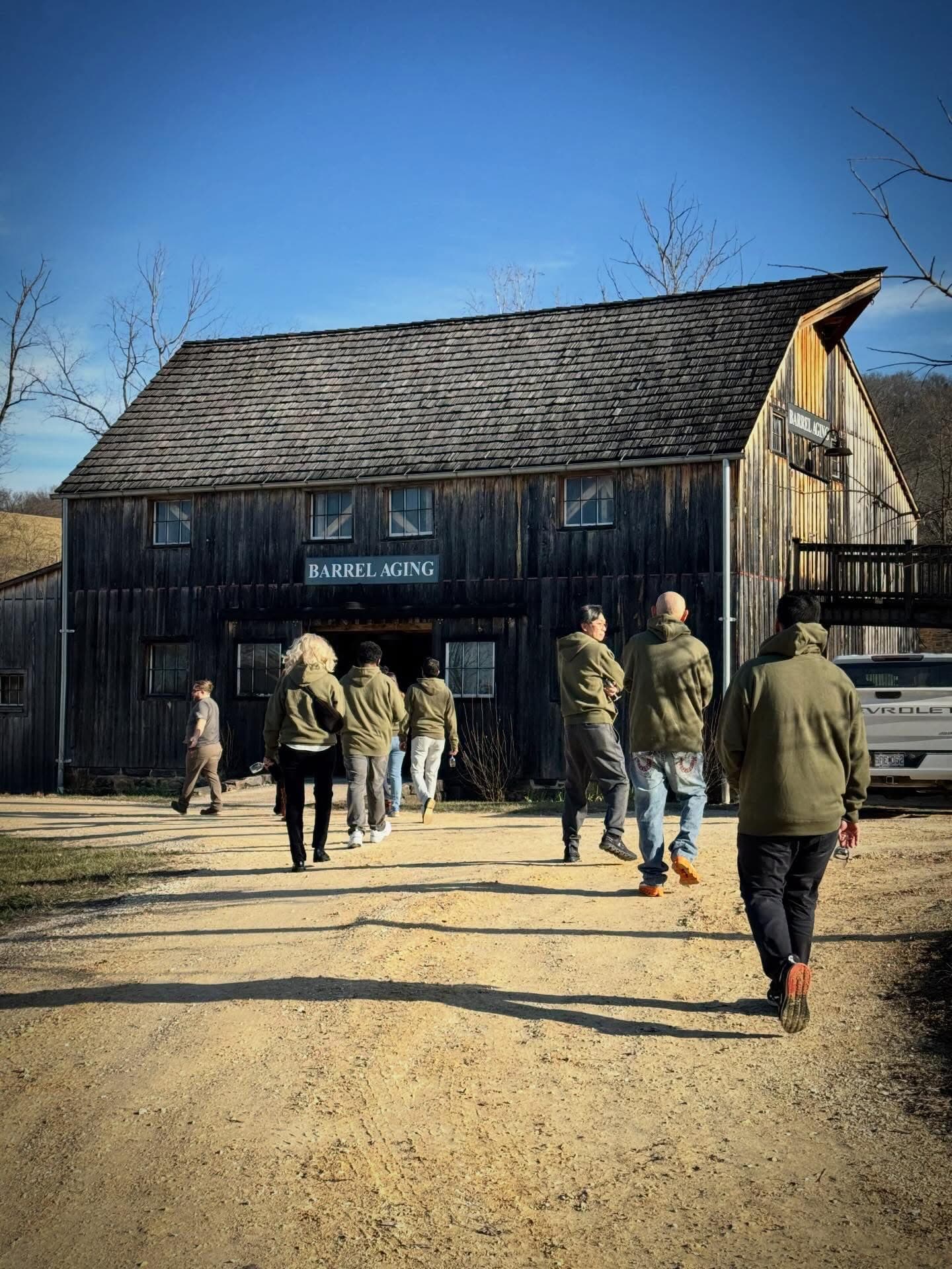 A group of people walks towards a barn labeled "BARREL AGING" on a sunny day.