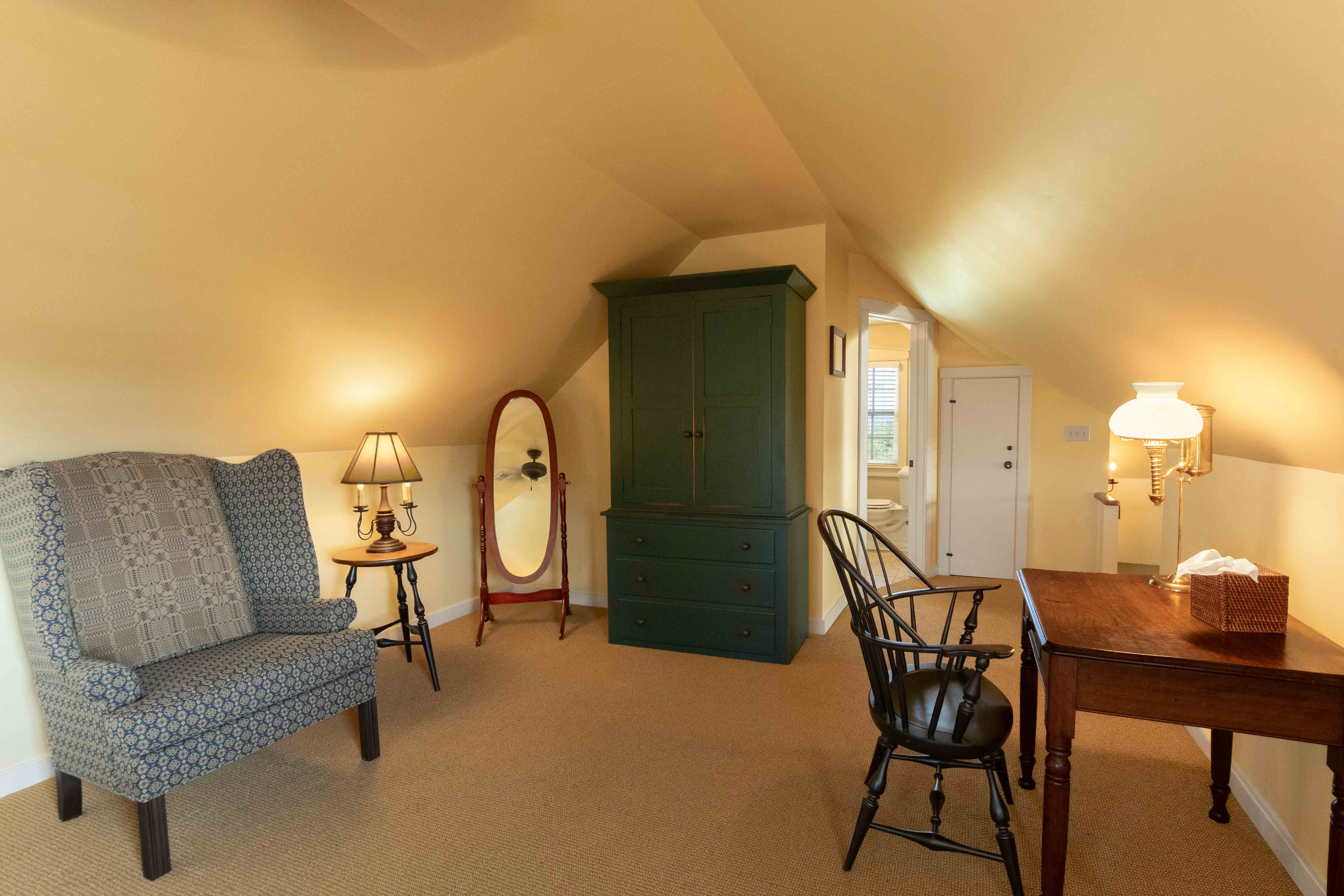 Cozy attic room featuring a patterned chair, a green wardrobe, and a wooden desk with a lamp.