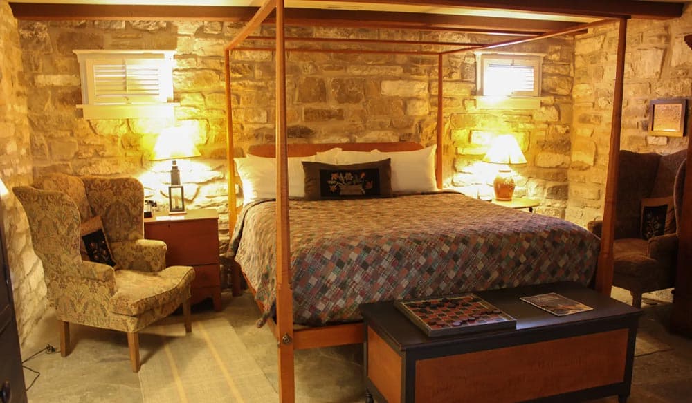 A cozy, rustic bedroom featuring a four-poster bed, armchairs, and stone walls.