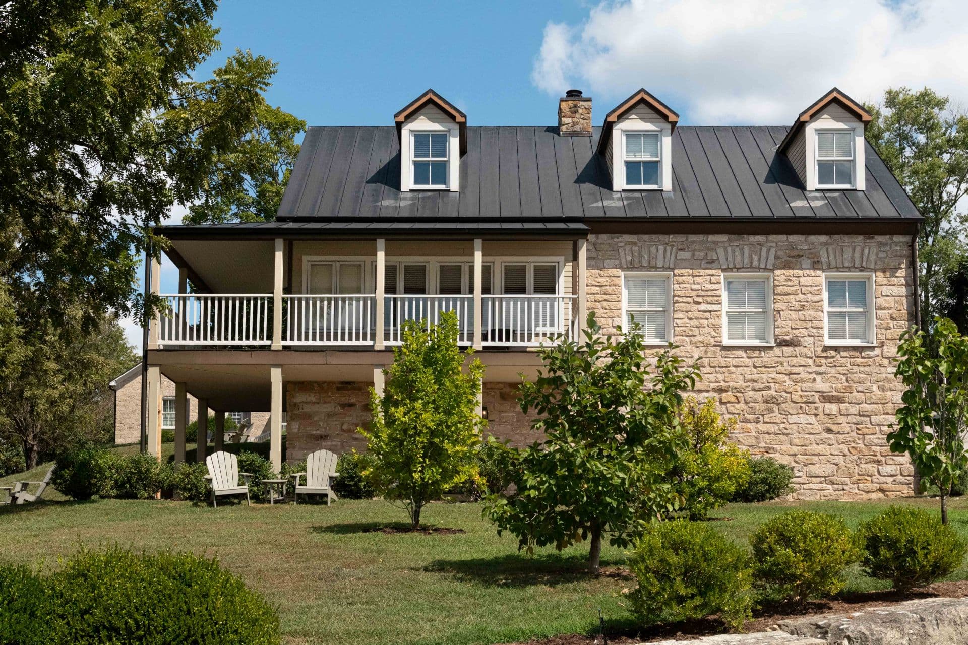 A modern stone house with a metal roof, featuring a front porch and landscaped yard.