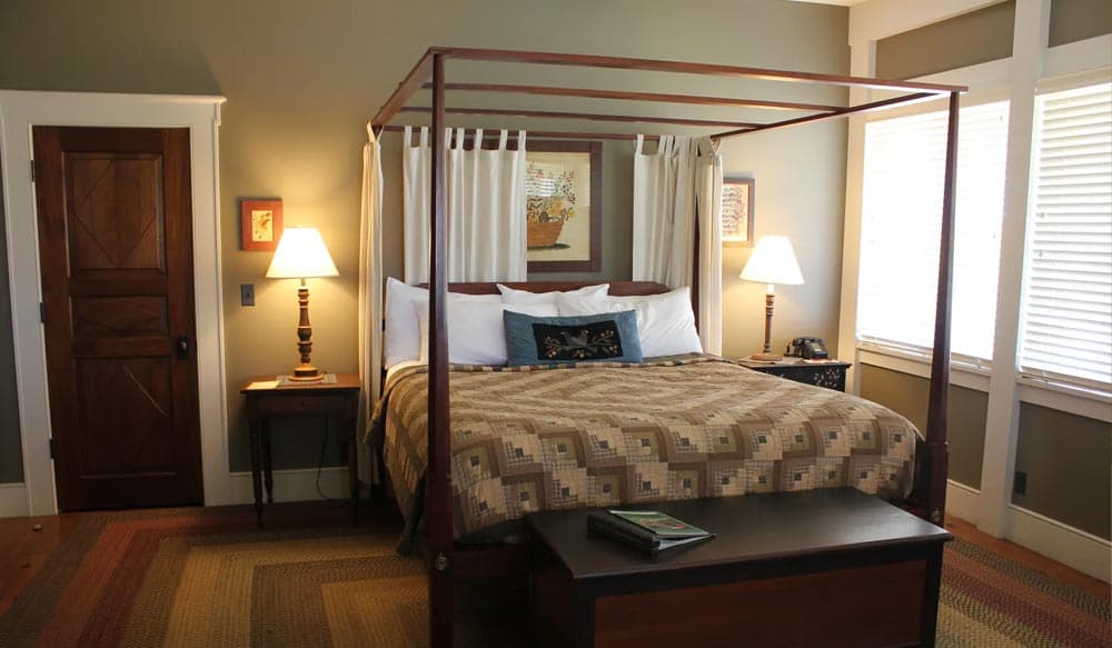 A cozy bedroom featuring a four-poster bed, bedside lamps, and natural light from window blinds.
