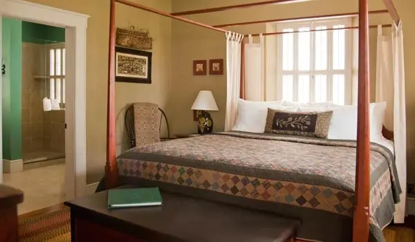 A cozy bedroom featuring a four-poster bed, a chair, a lamp, and an adjoining bathroom.