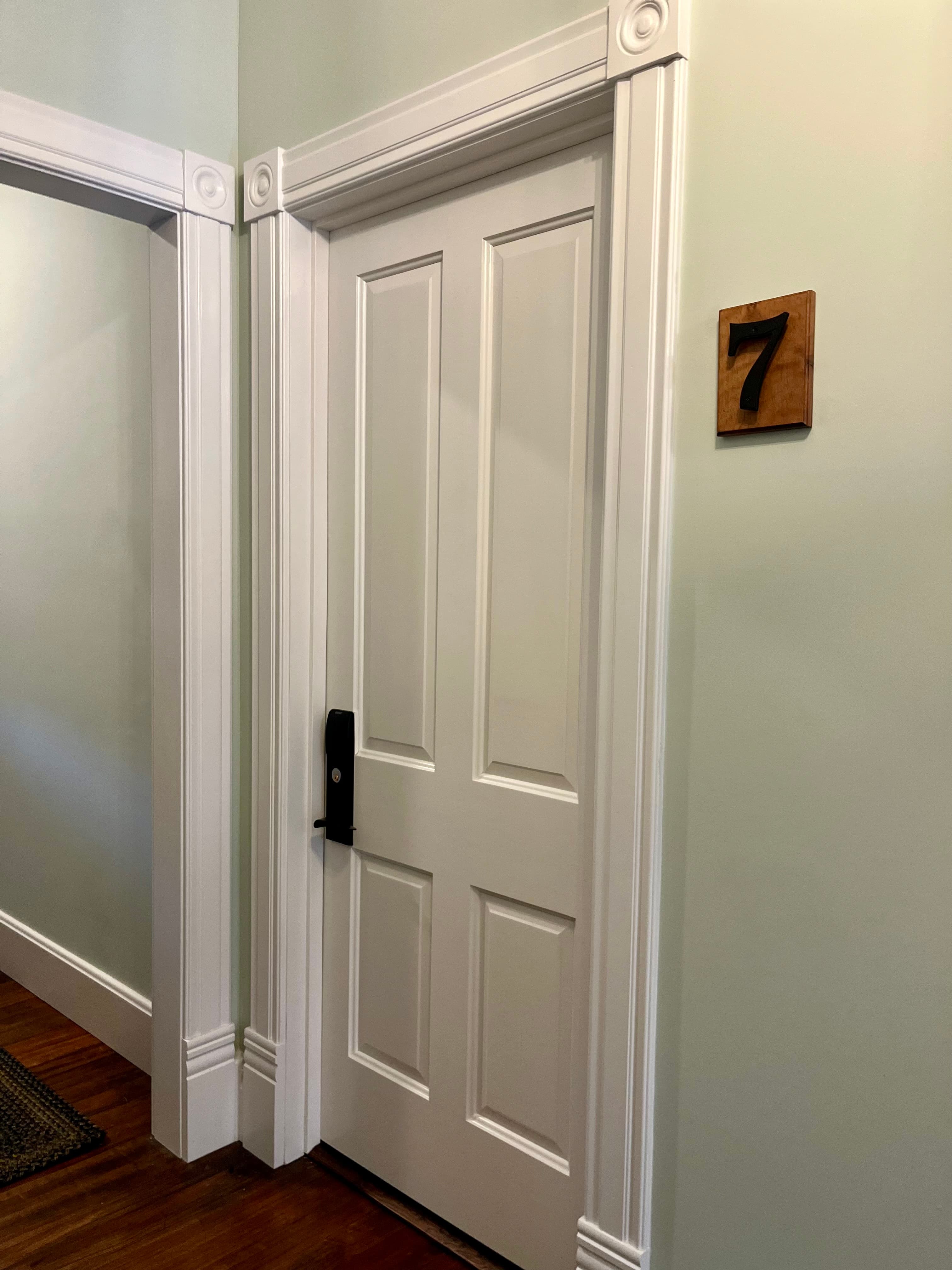 A white door with a black handle and the number 7 displayed on a wooden plaque is set against a light green wall.