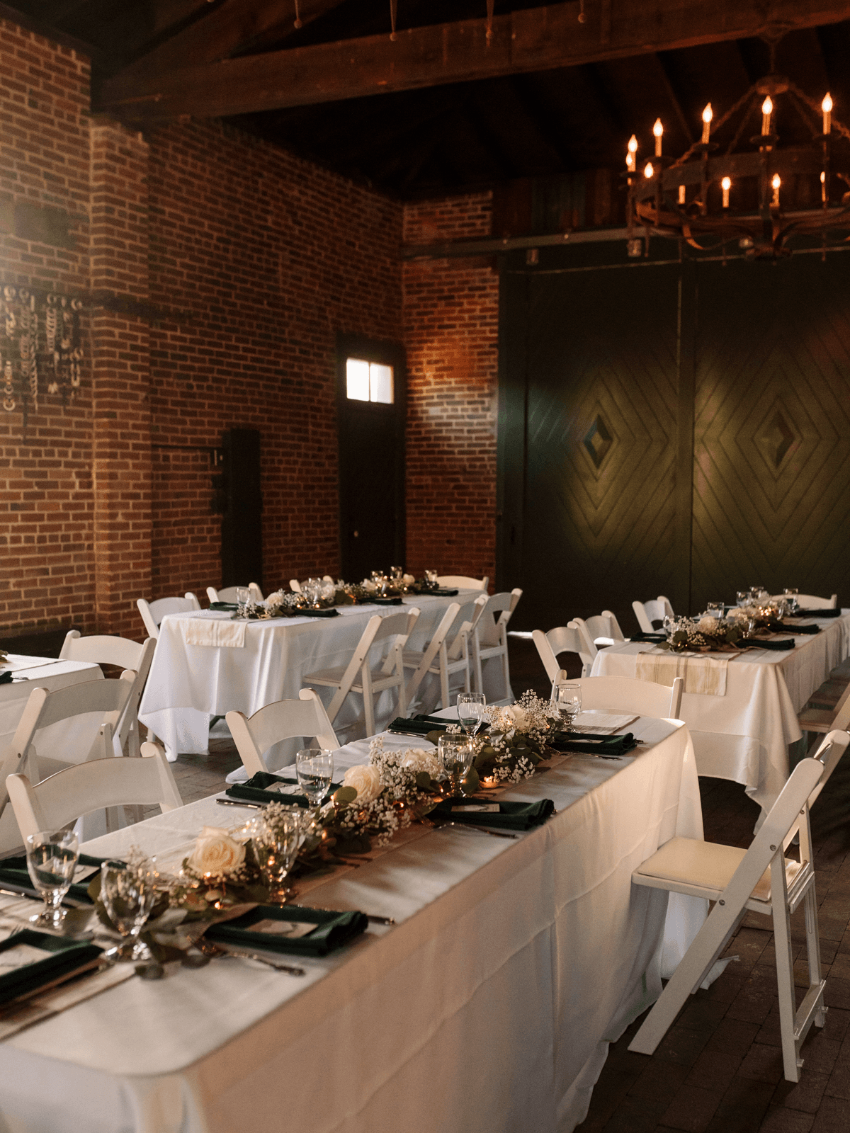 A beautifully arranged dining setup with white tables, greenery, and floral decorations in a rustic brick venue.