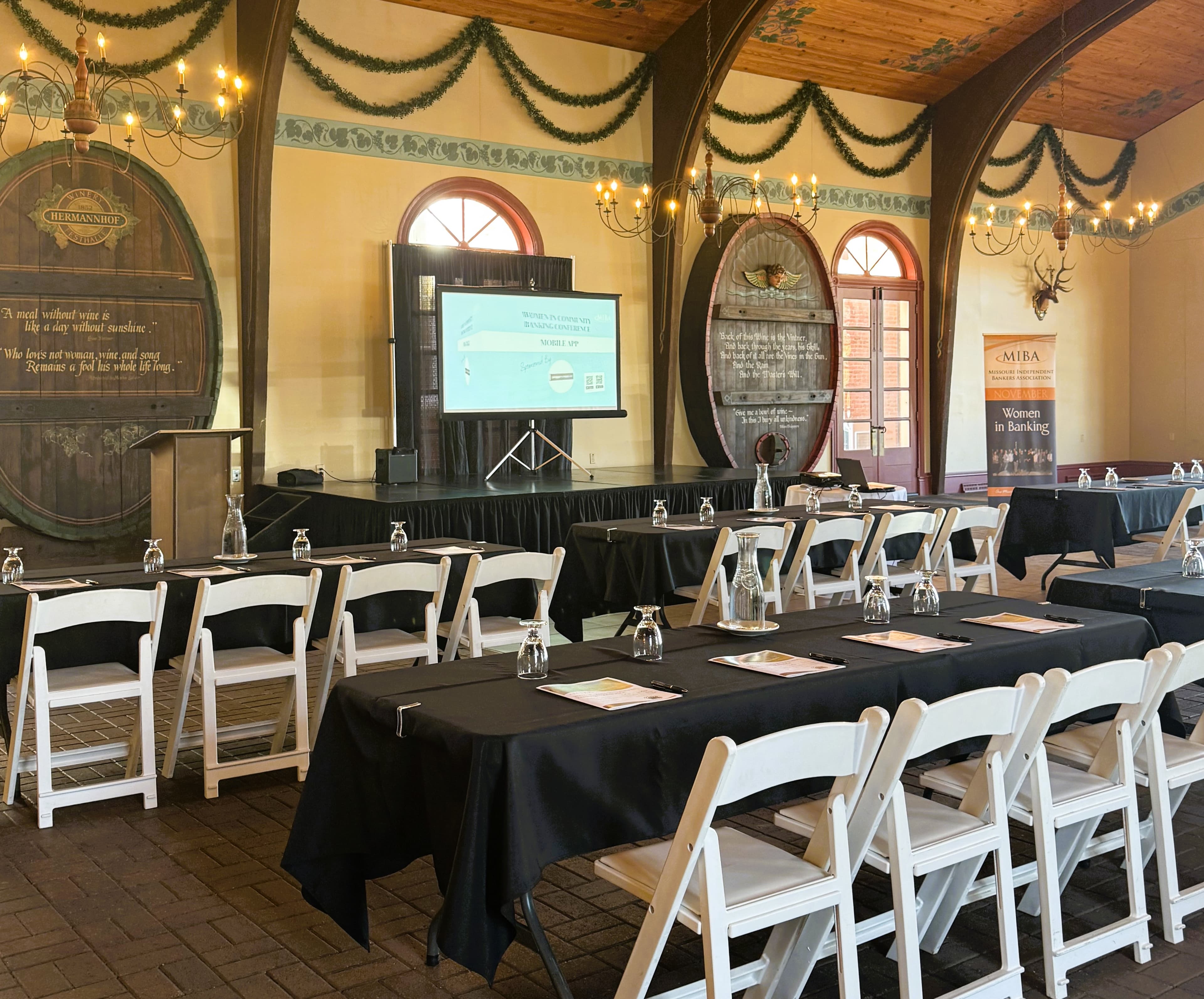 A spacious event room set up with tables and chairs for a presentation, featuring a large screen and decorative wall elements.