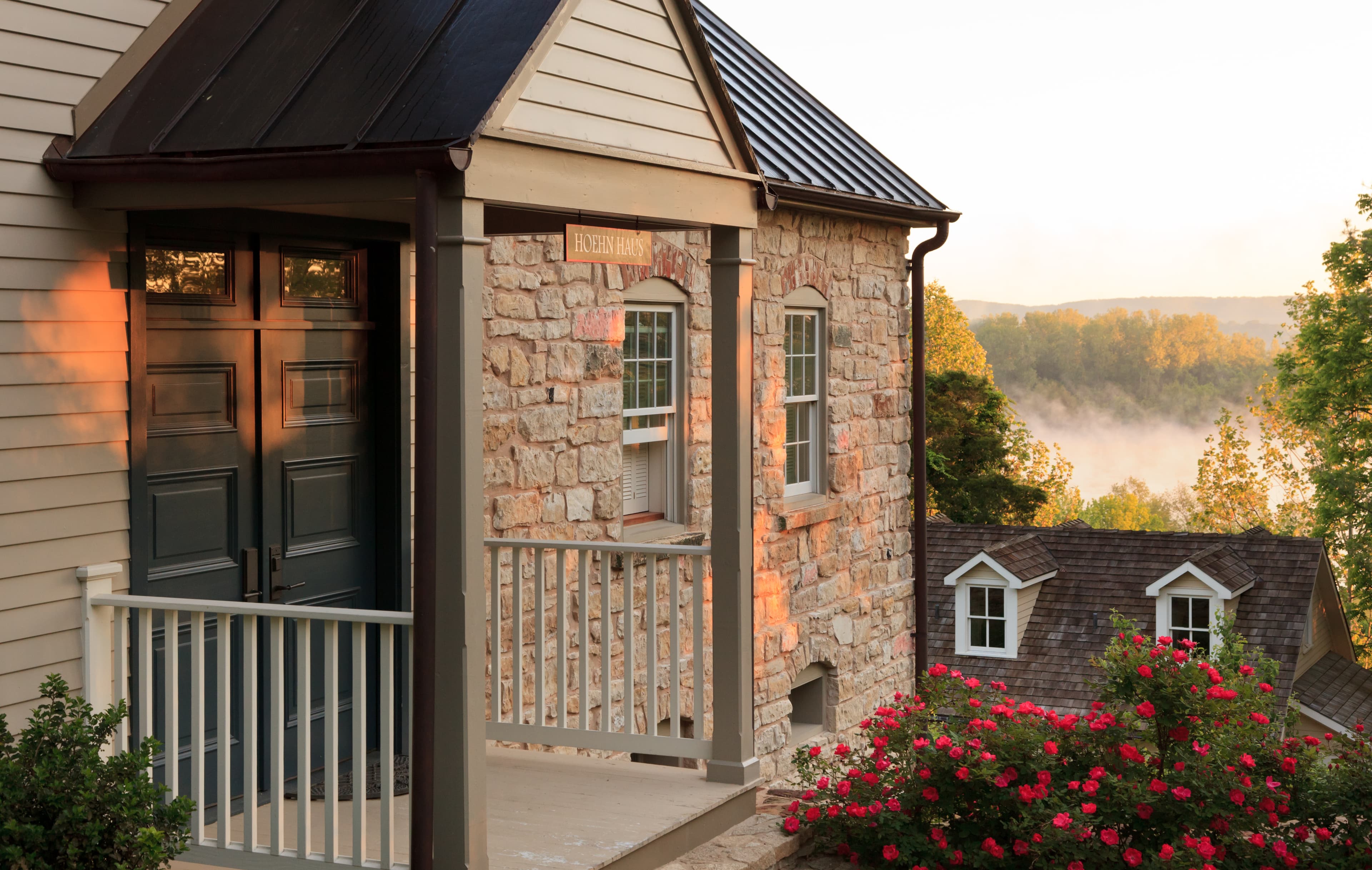A stone house entrance with a porch, overlooking a misty landscape and surrounded by blooming flowers.