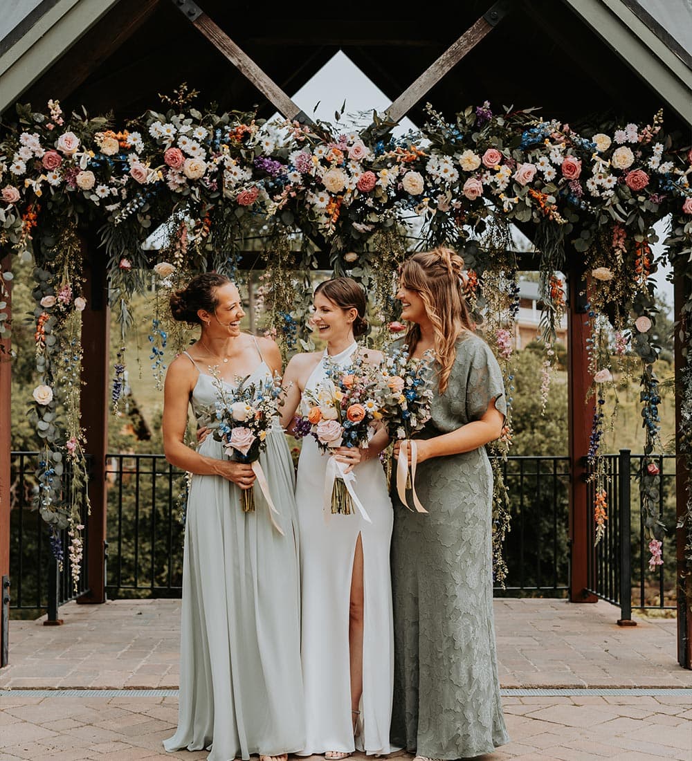 Three smiling women in elegant dresses stand together under a floral arch, each holding a bouquet.