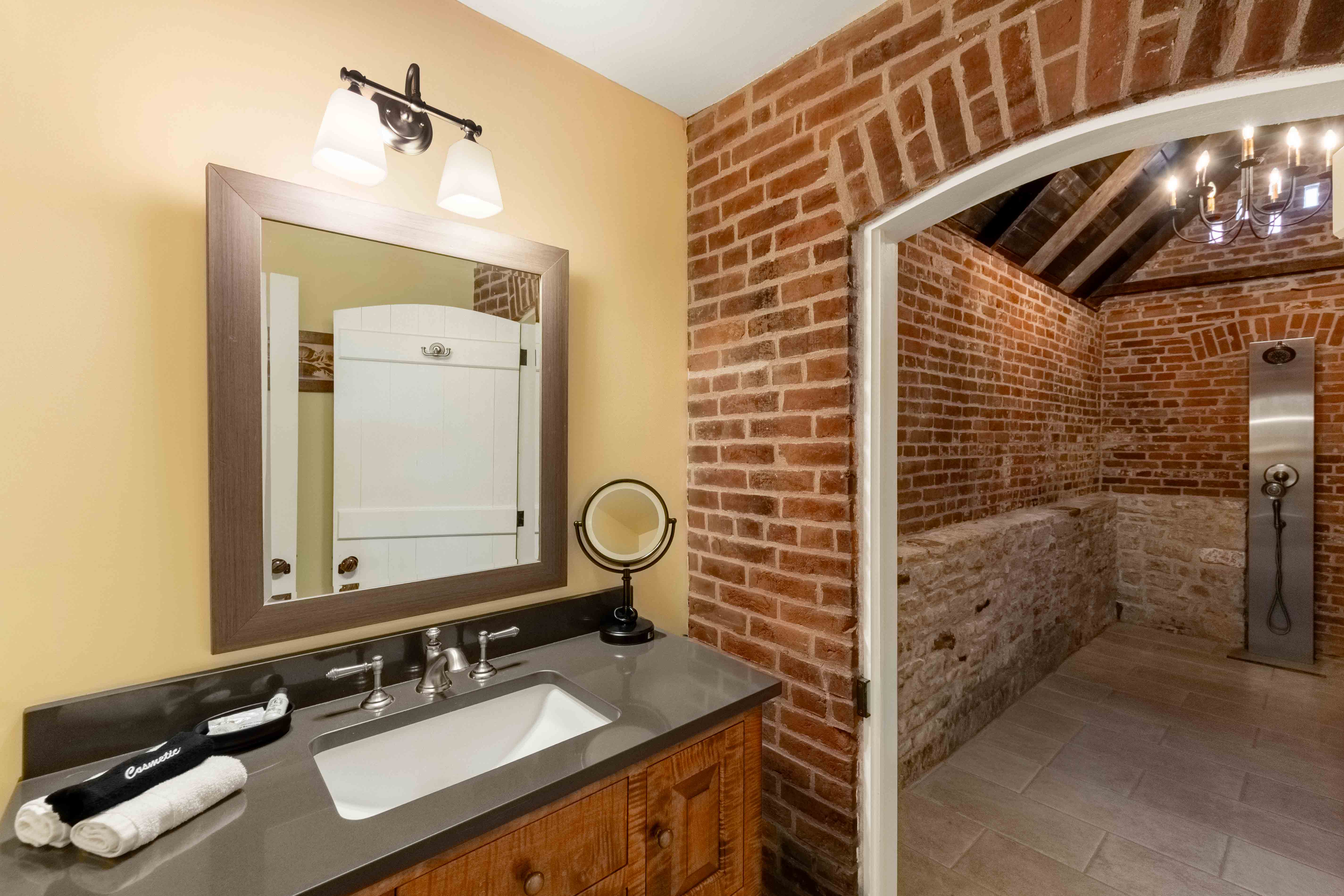 A bathroom featuring a wooden vanity with a mirror and sink, brick walls, and an arched doorway leading to a rustic shower area.