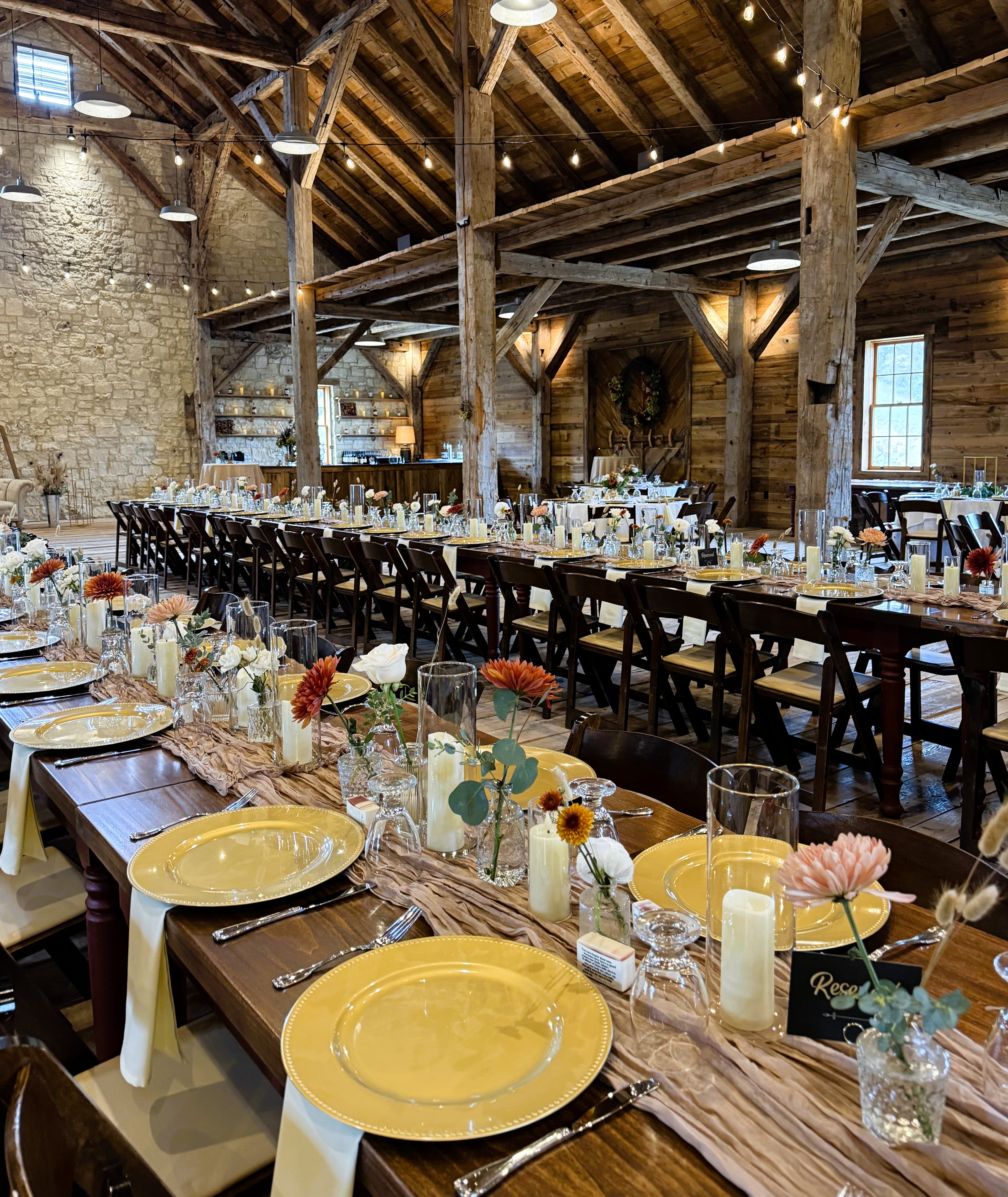 A rustic dining setup featuring long tables adorned with yellow plates, glassware, and floral centerpieces in a warm wooden barn.
