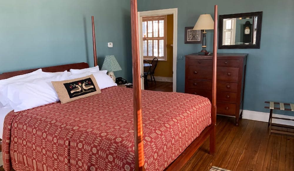 A cozy bedroom featuring a four-poster bed with a red patterned coverlet and a wooden dresser against a blue wall.