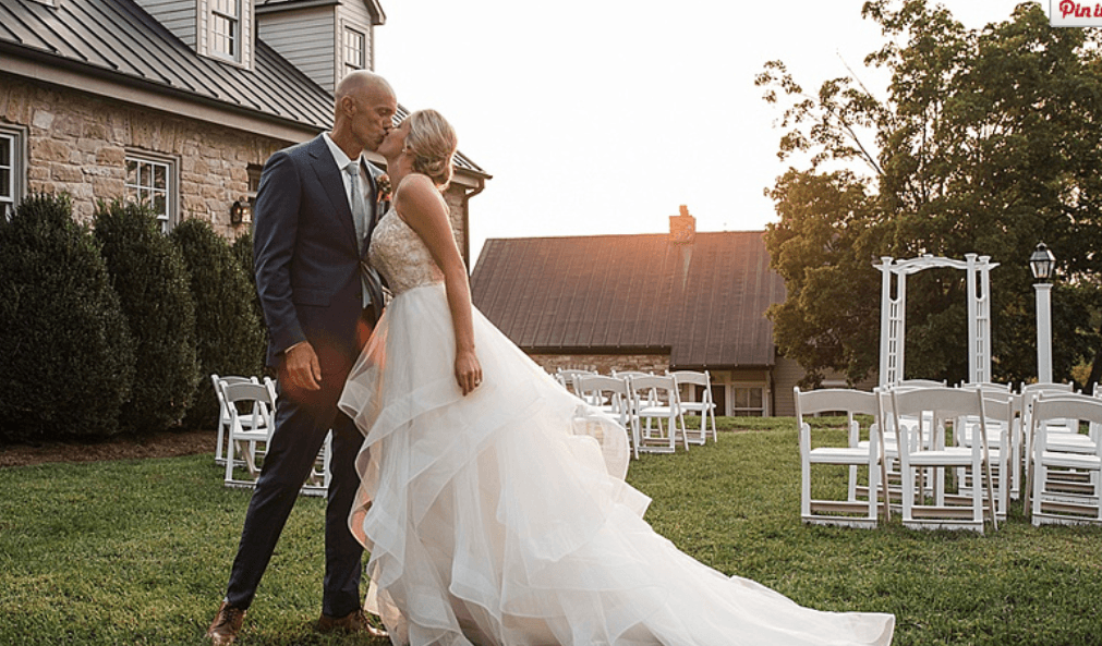 A couple shares a kiss outdoors at sunset, with empty chairs arranged for a wedding ceremony in the background.