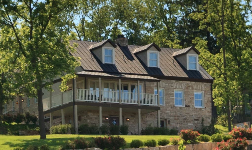A two-story stone house with a metal roof and a front porch surrounded by greenery.