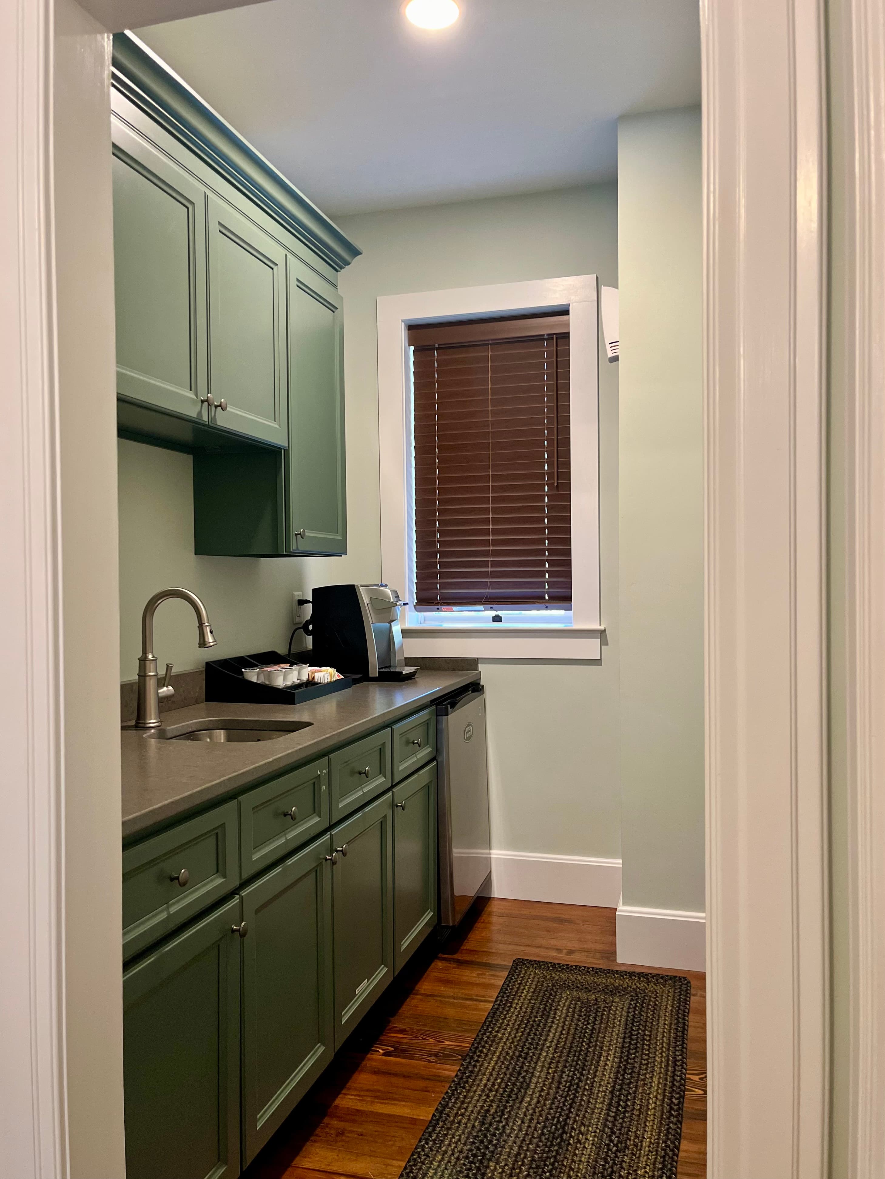 A narrow kitchen area featuring green cabinets, a stainless steel sink, and a window with brown blinds.