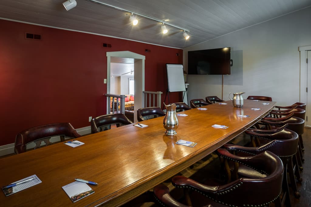 A large wooden conference table surrounded by chairs in a meeting room with red walls and a TV.