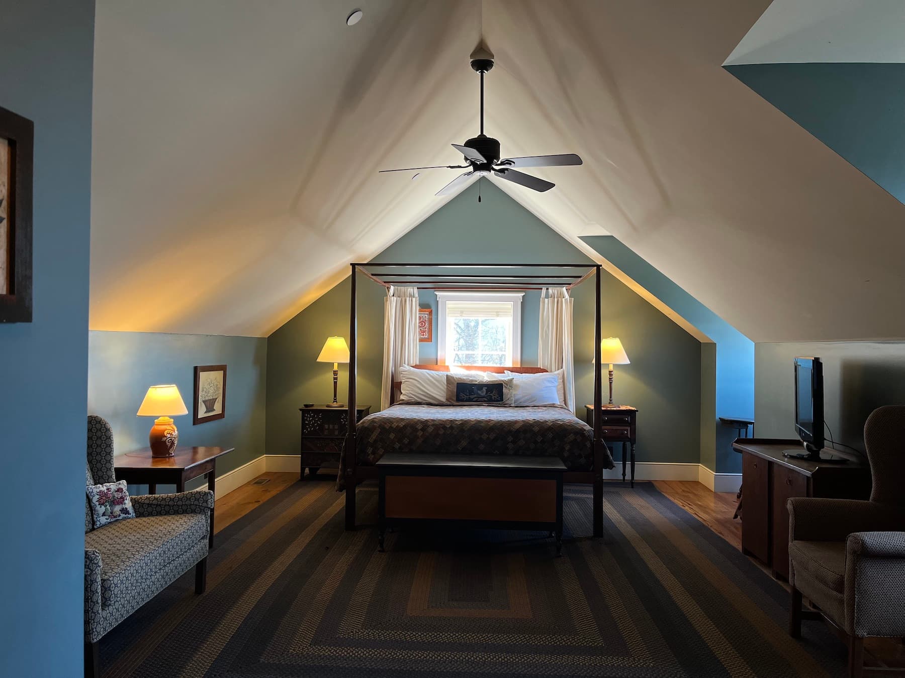 A cozy, well-lit attic bedroom featuring a large bed, armchairs, and a ceiling fan.