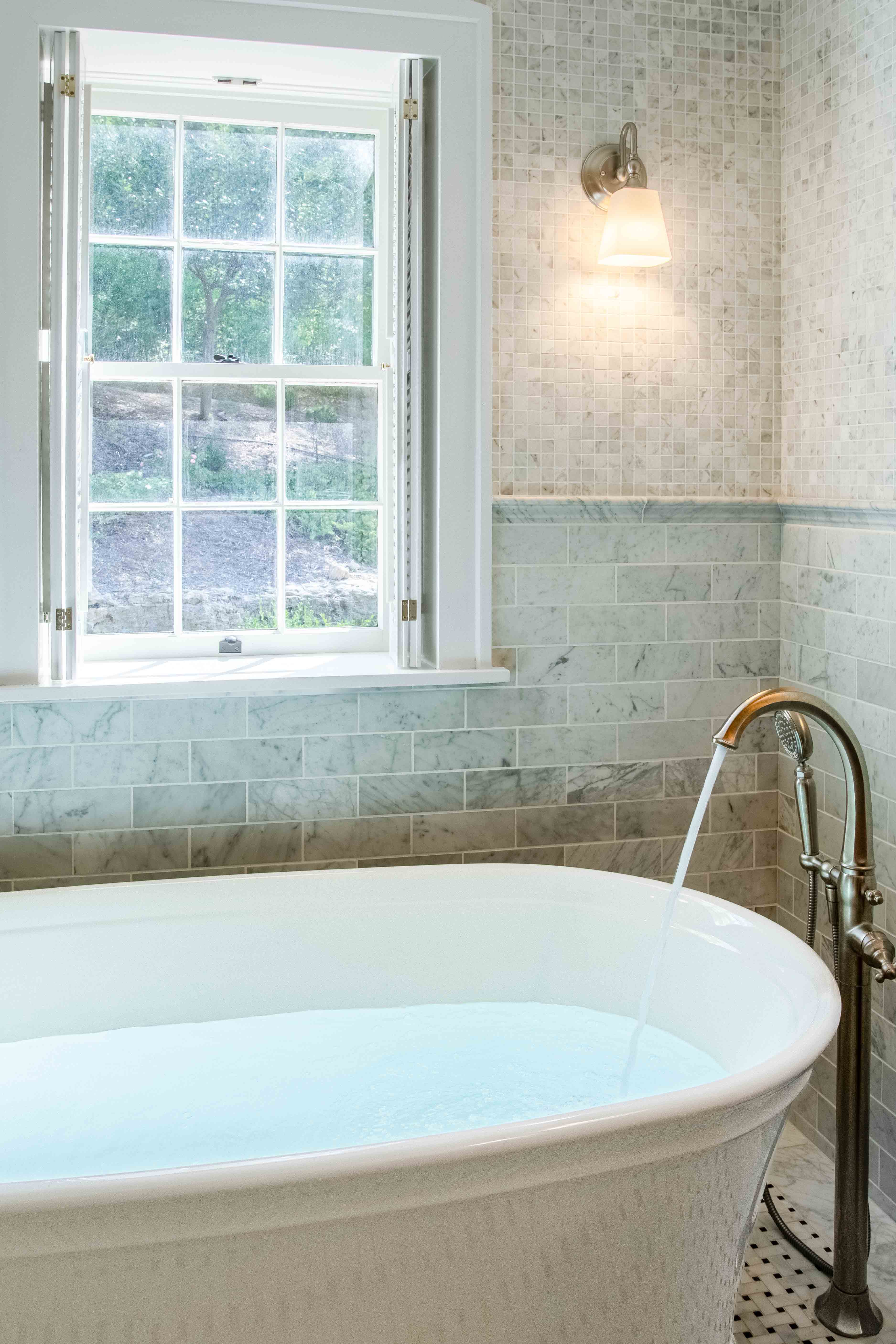 A white freestanding bathtub with running water beneath a window in a tiled bathroom.