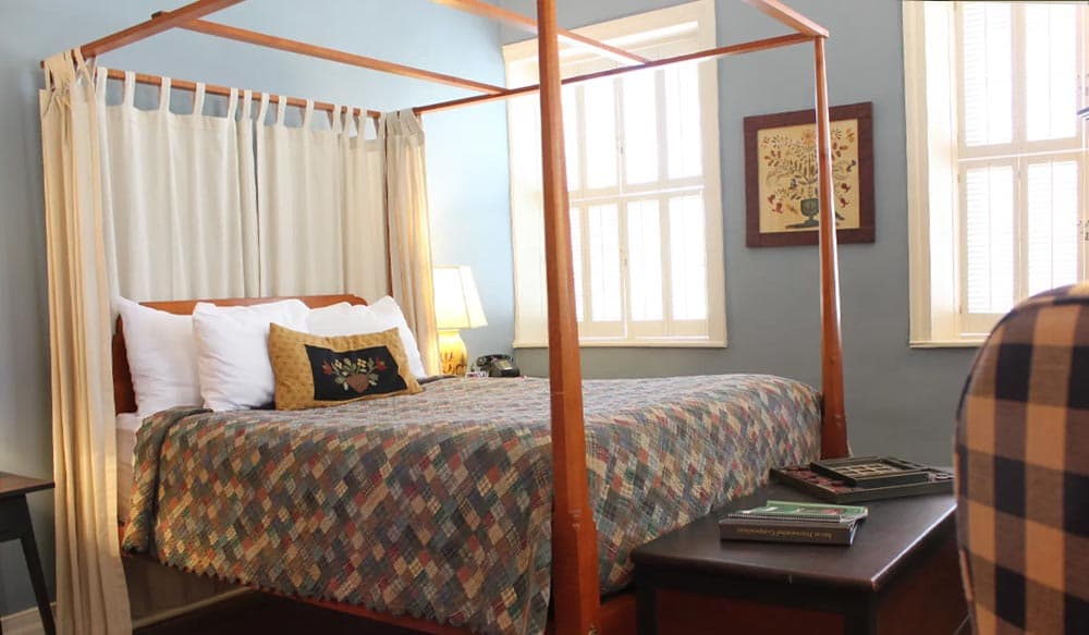 A cozy bedroom featuring a four-poster bed with a colorful quilt and windows letting in natural light.