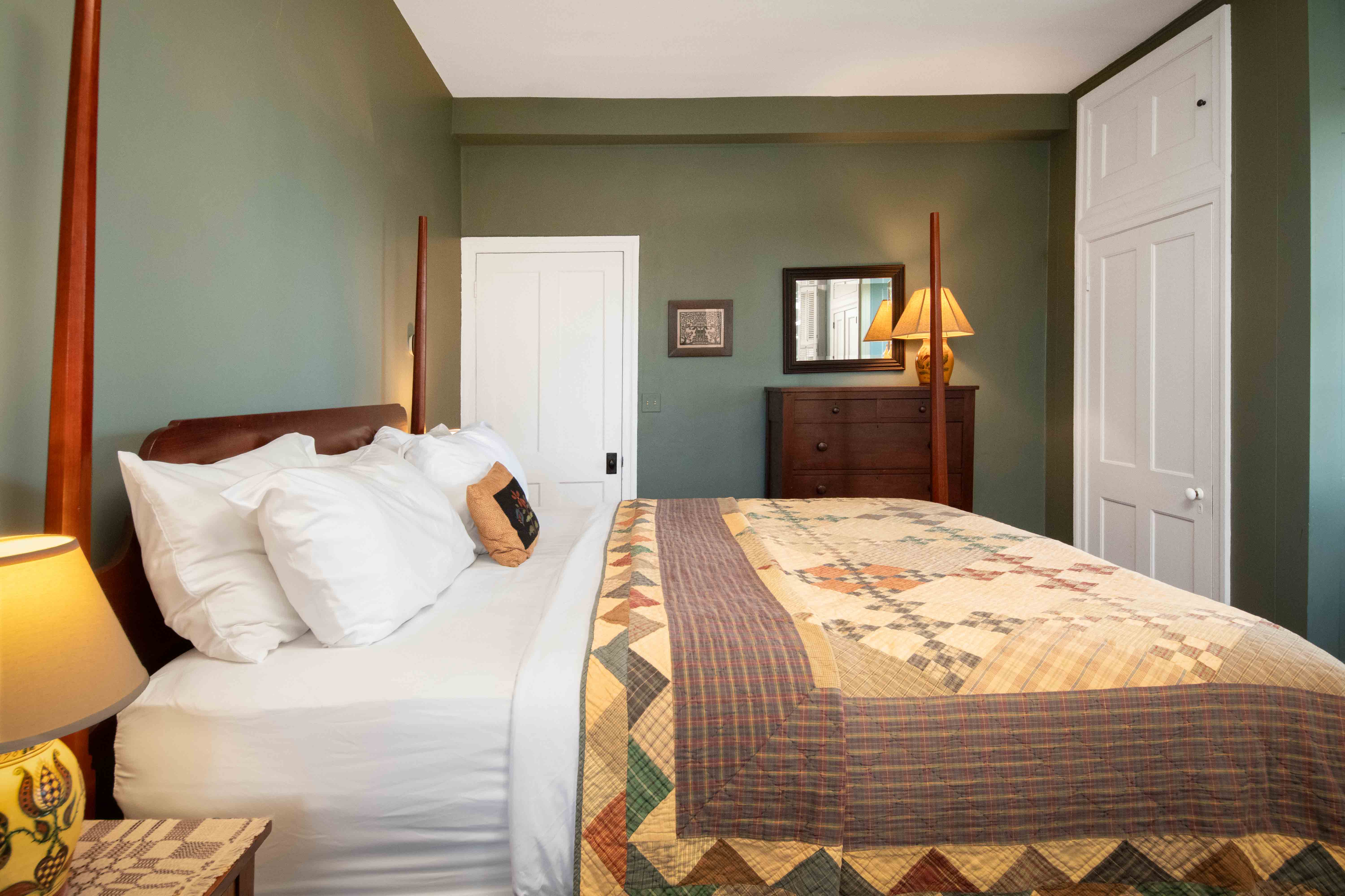 A cozy bedroom featuring a bed with a patterned quilt, a bedside lamp, and a wooden dresser against a green wall.