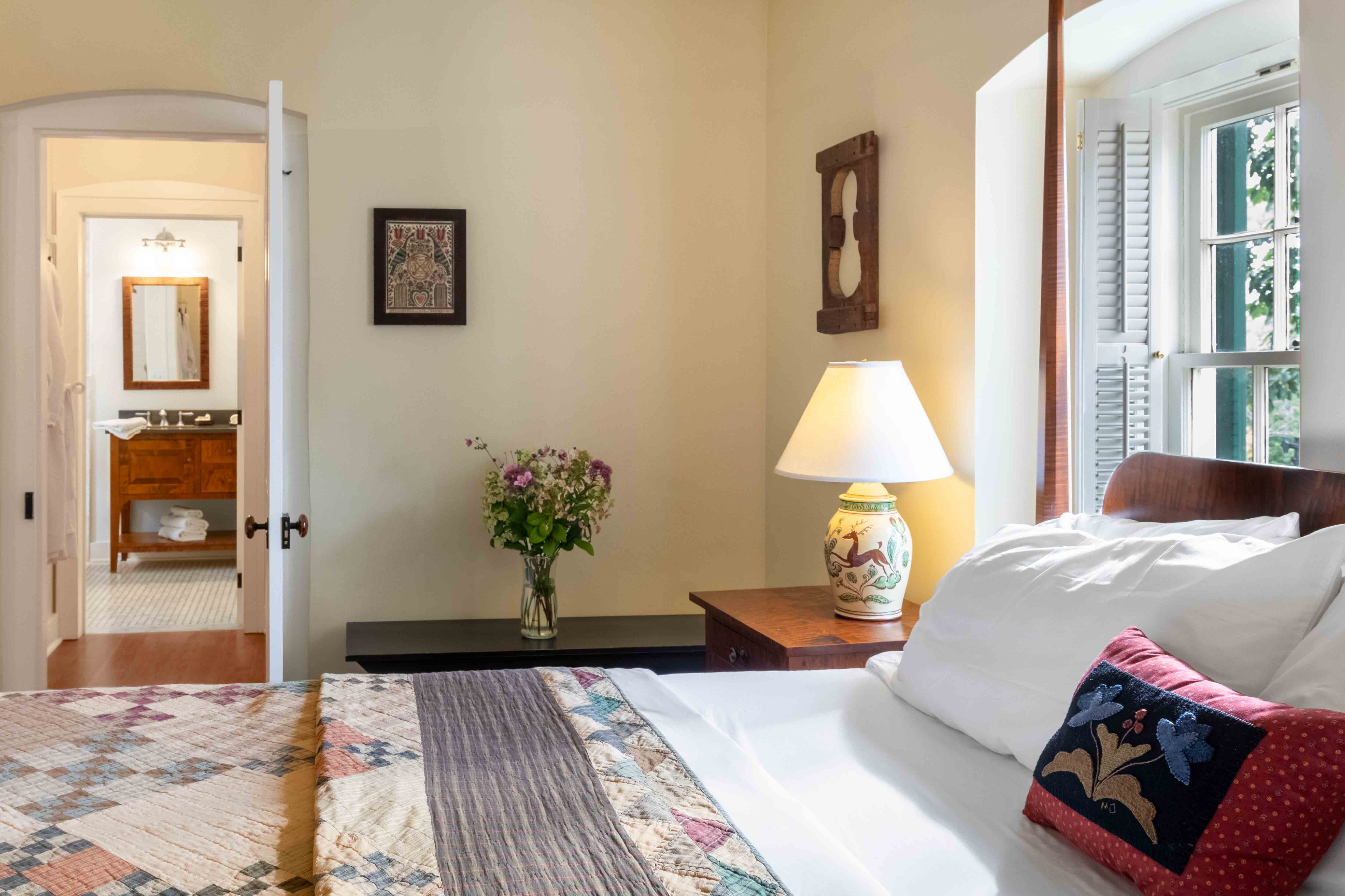 A cozy bedroom featuring a quilted bed, a lamp, and an open doorway leading to a bathroom.