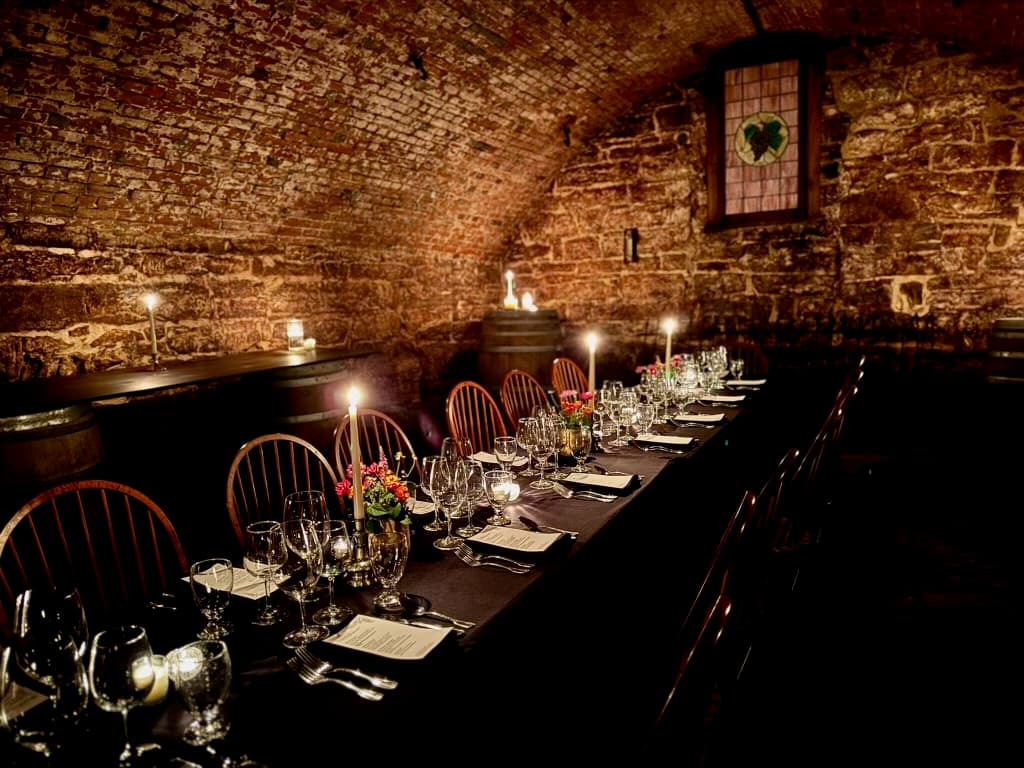A dimly lit cellar with a long dining table set for a formal meal, adorned with candles and flowers.