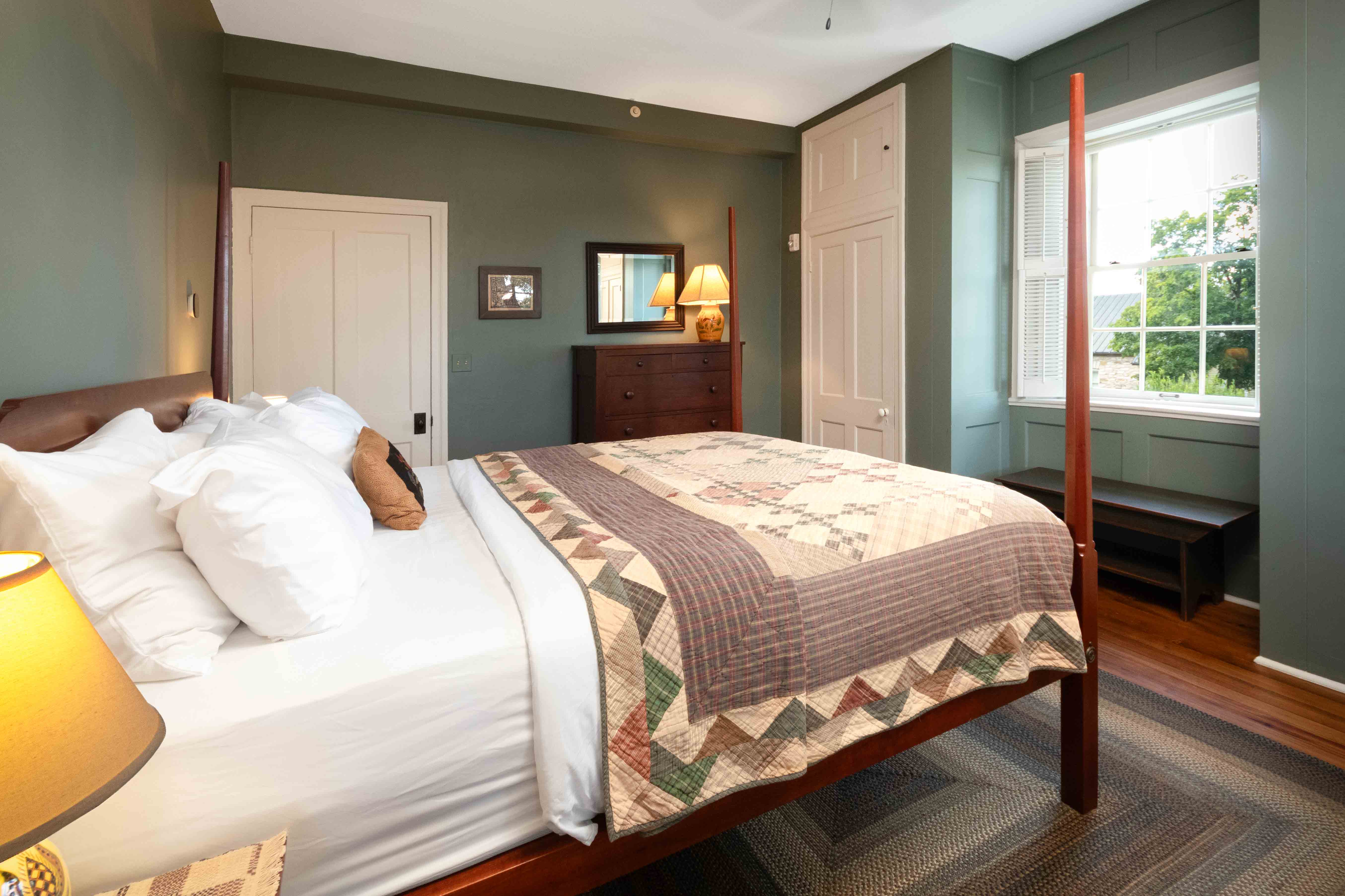 A cozy bedroom with a quilted bed, wooden furniture, and green walls, featuring natural light from a window.