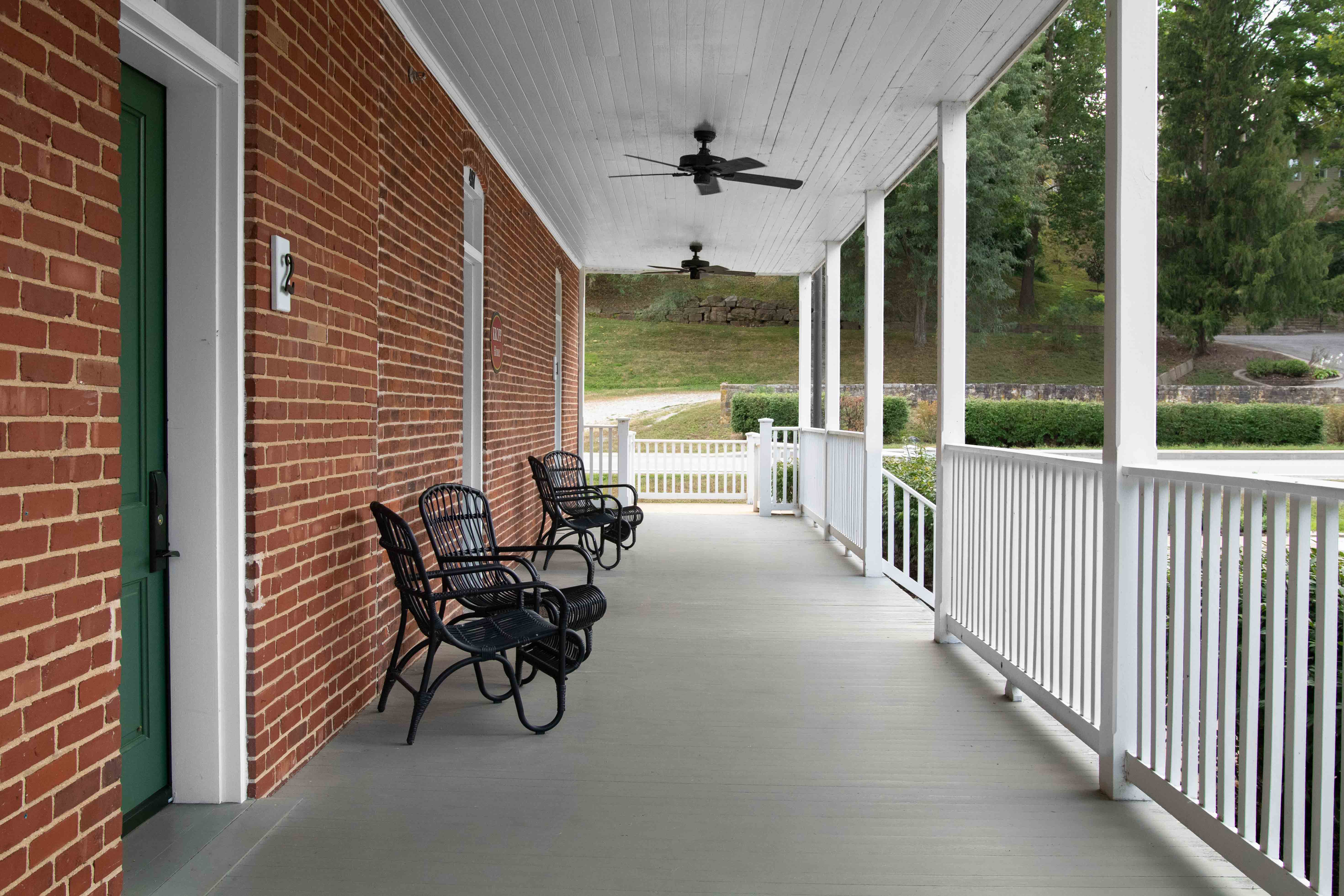 A covered porch with black chairs and a ceiling fan, surrounded by greenery.