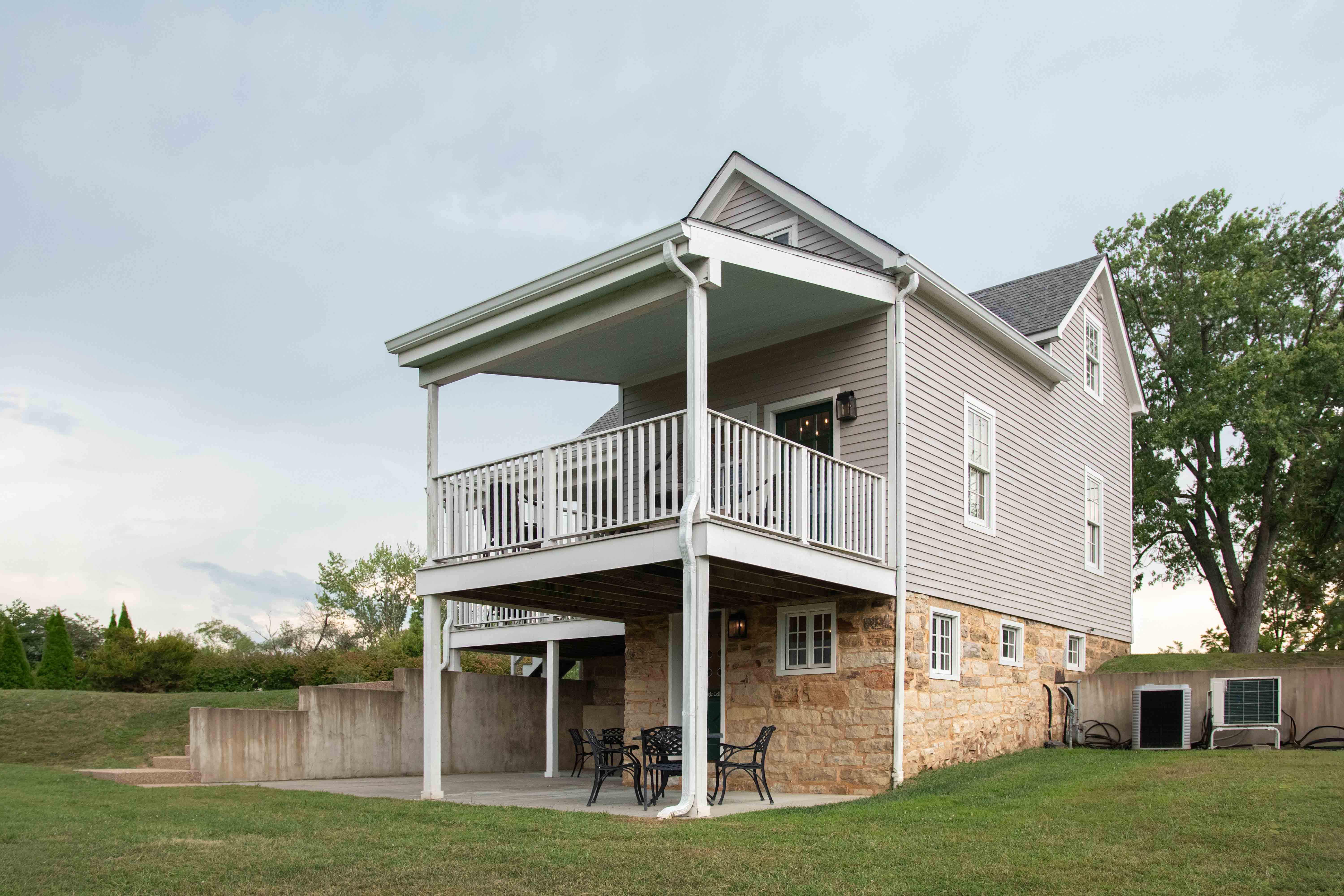 A two-story house with a stone foundation and a covered porch, surrounded by greenery.