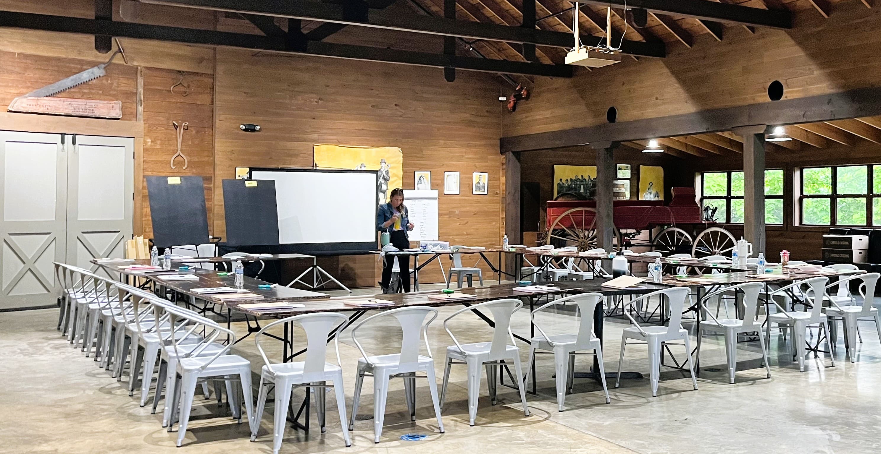 A spacious indoor room set up for an event, featuring multiple tables, chairs, and a presentation screen.