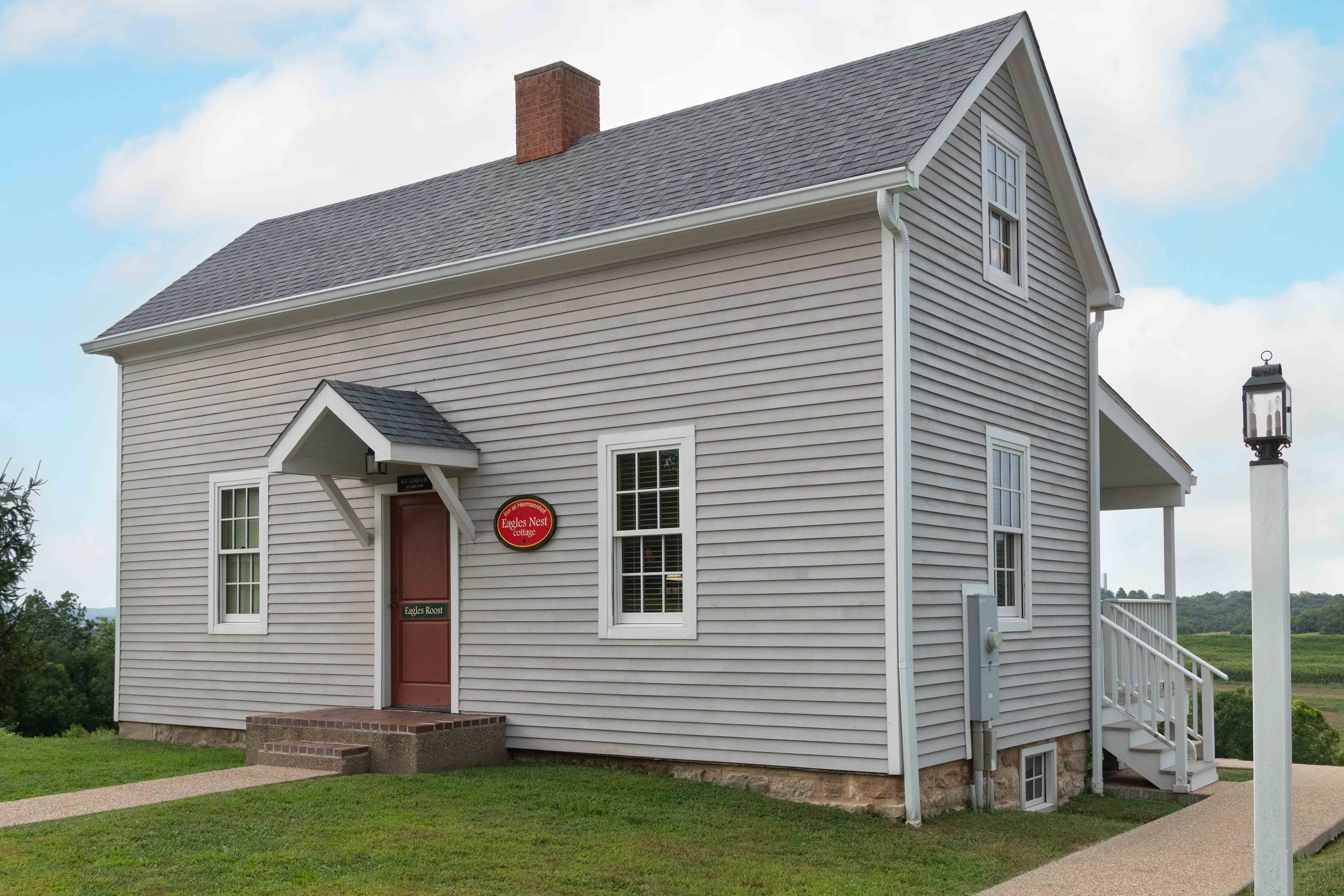 A light gray house with a red door and sign, set in a grassy area under a cloudy sky.