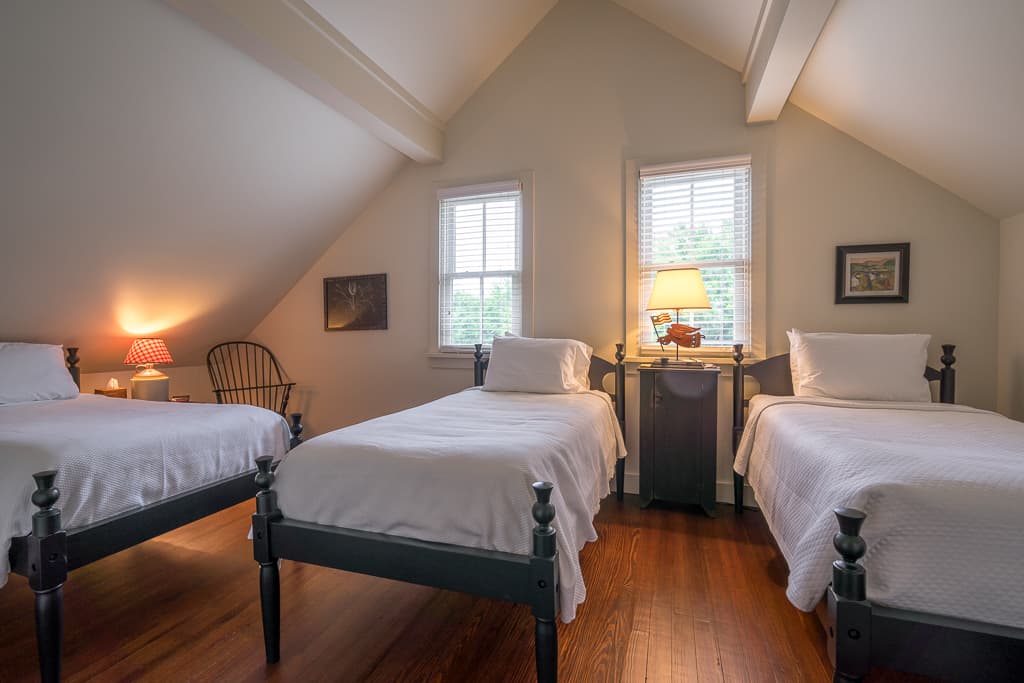 A cozy attic bedroom featuring three twin beds, a rocking chair, and warm lighting from table lamps.