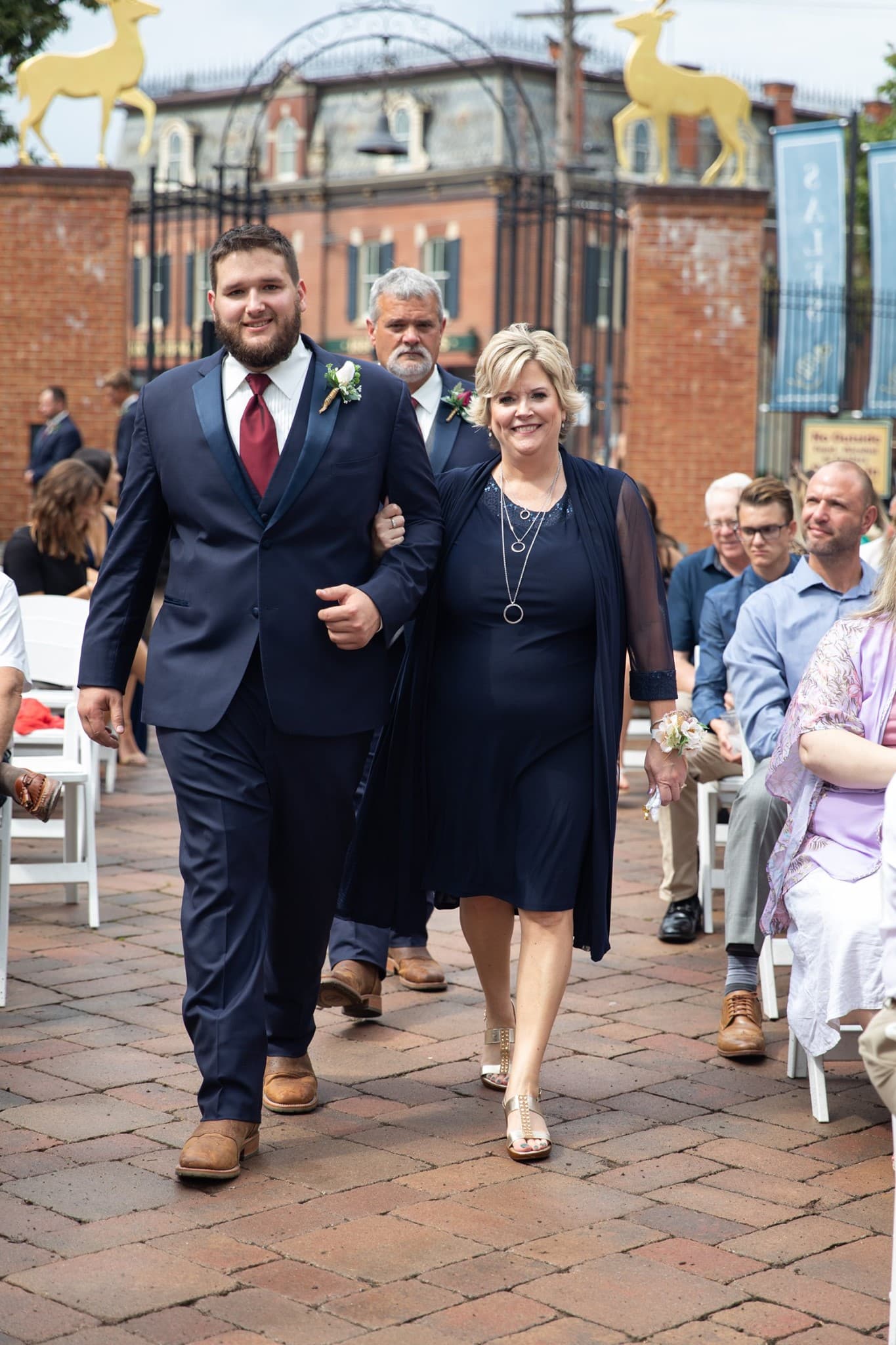 A man in a navy suit walks arm-in-arm with a woman in a navy dress and shawl during a wedding procession.