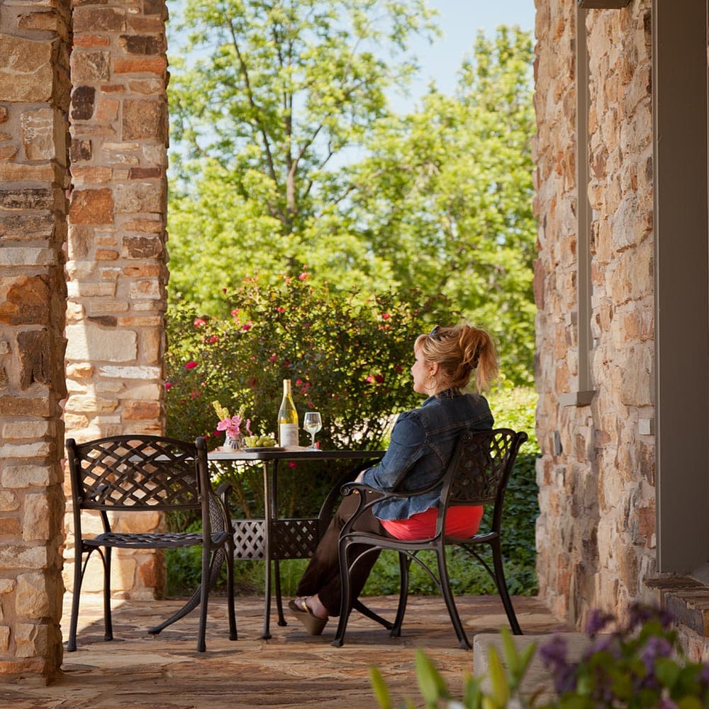 A woman sits at a table on a patio, enjoying wine and surrounded by greenery.
