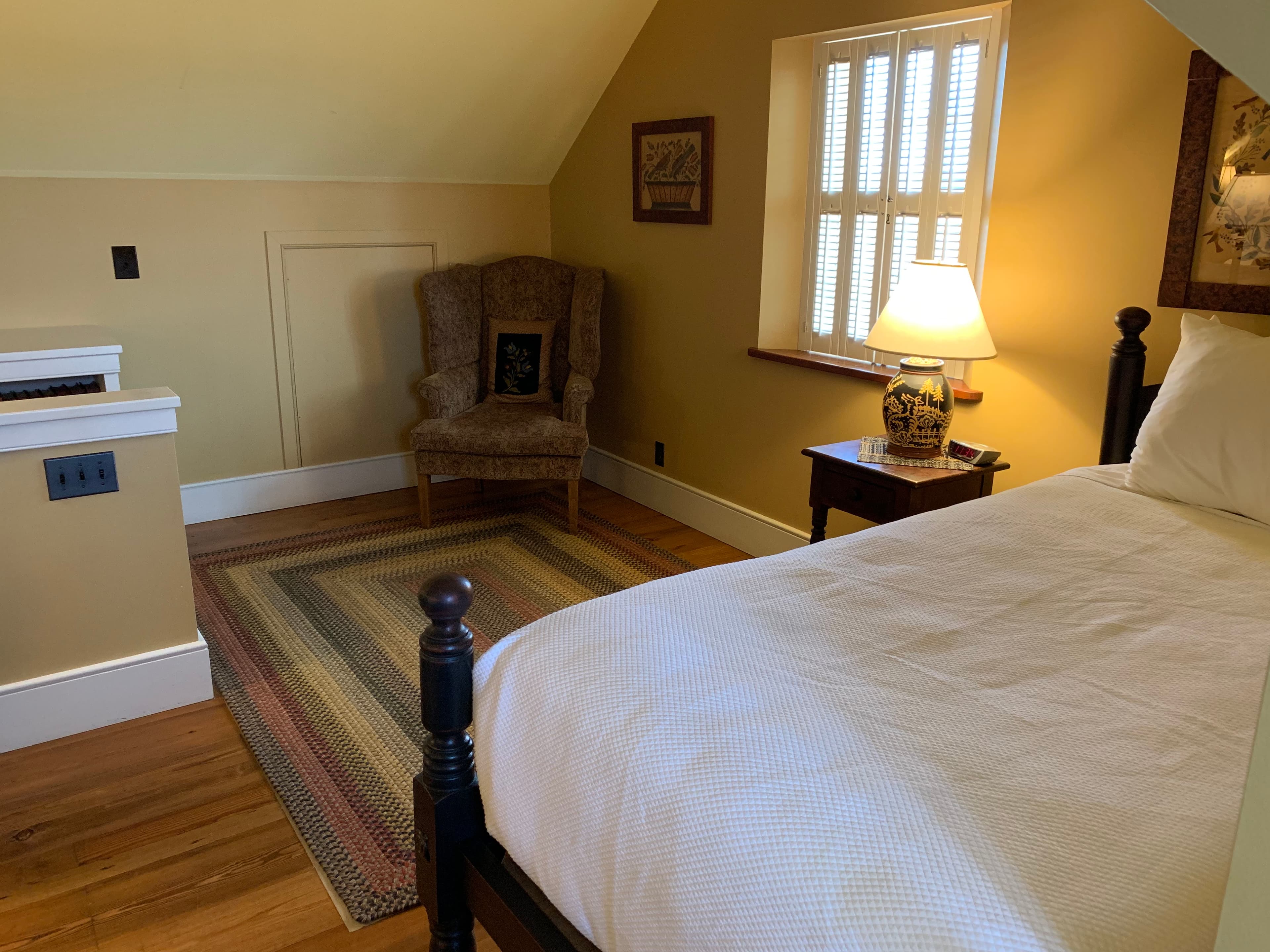 A cozy bedroom featuring a bed, a patterned rug, a lamp, and a comfortable armchair by a window.