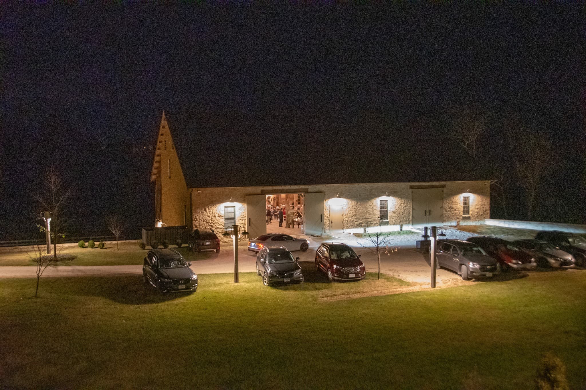 A stone building with a peaked roof is illuminated at night, surrounded by parked cars.