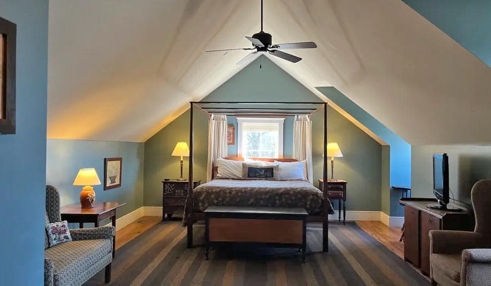 Cozy attic bedroom featuring a four-poster bed, armchairs, and a fan under a sloped ceiling.