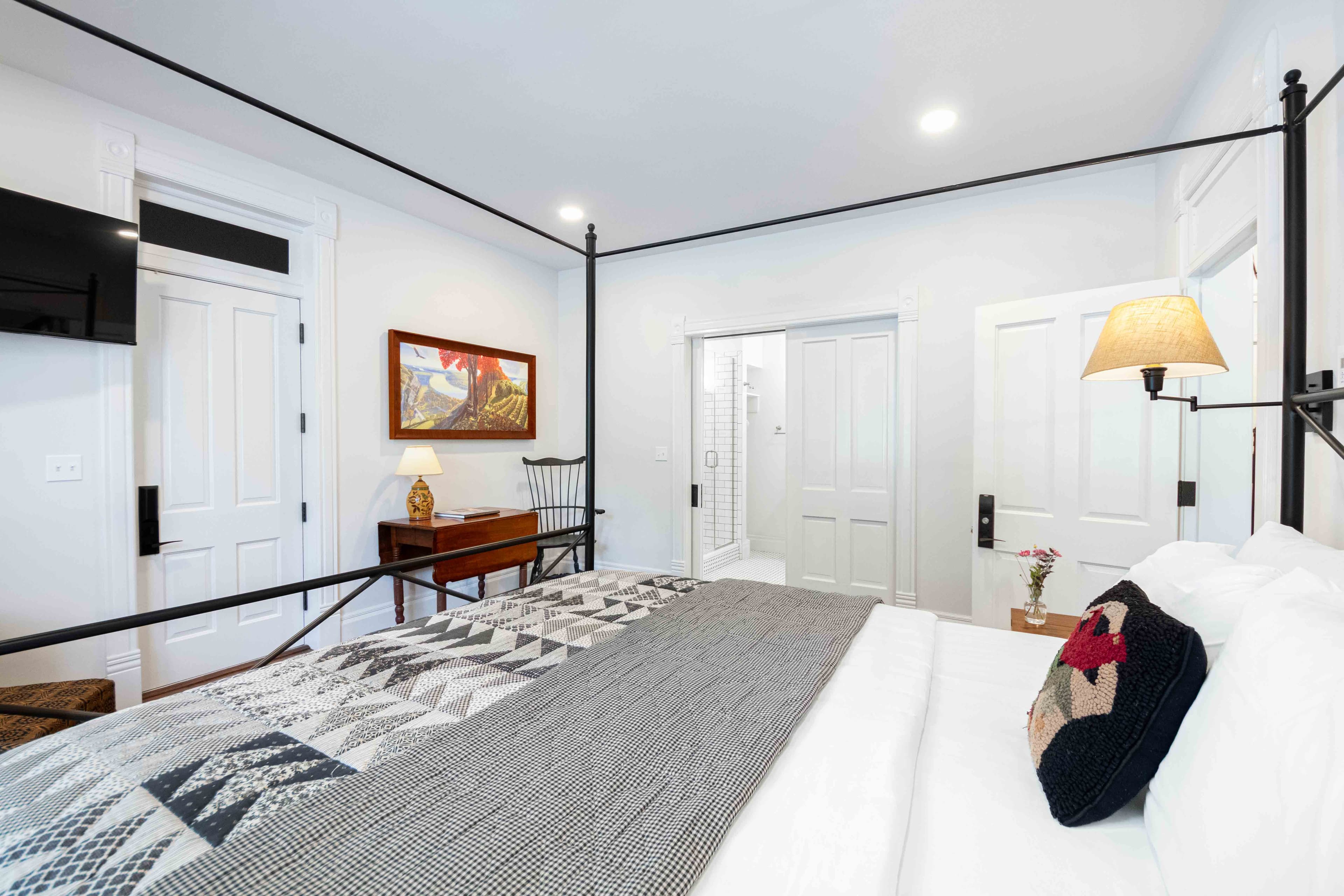 A cozy bedroom featuring a bed with a patterned quilt, a small table, and doors leading to a bathroom.