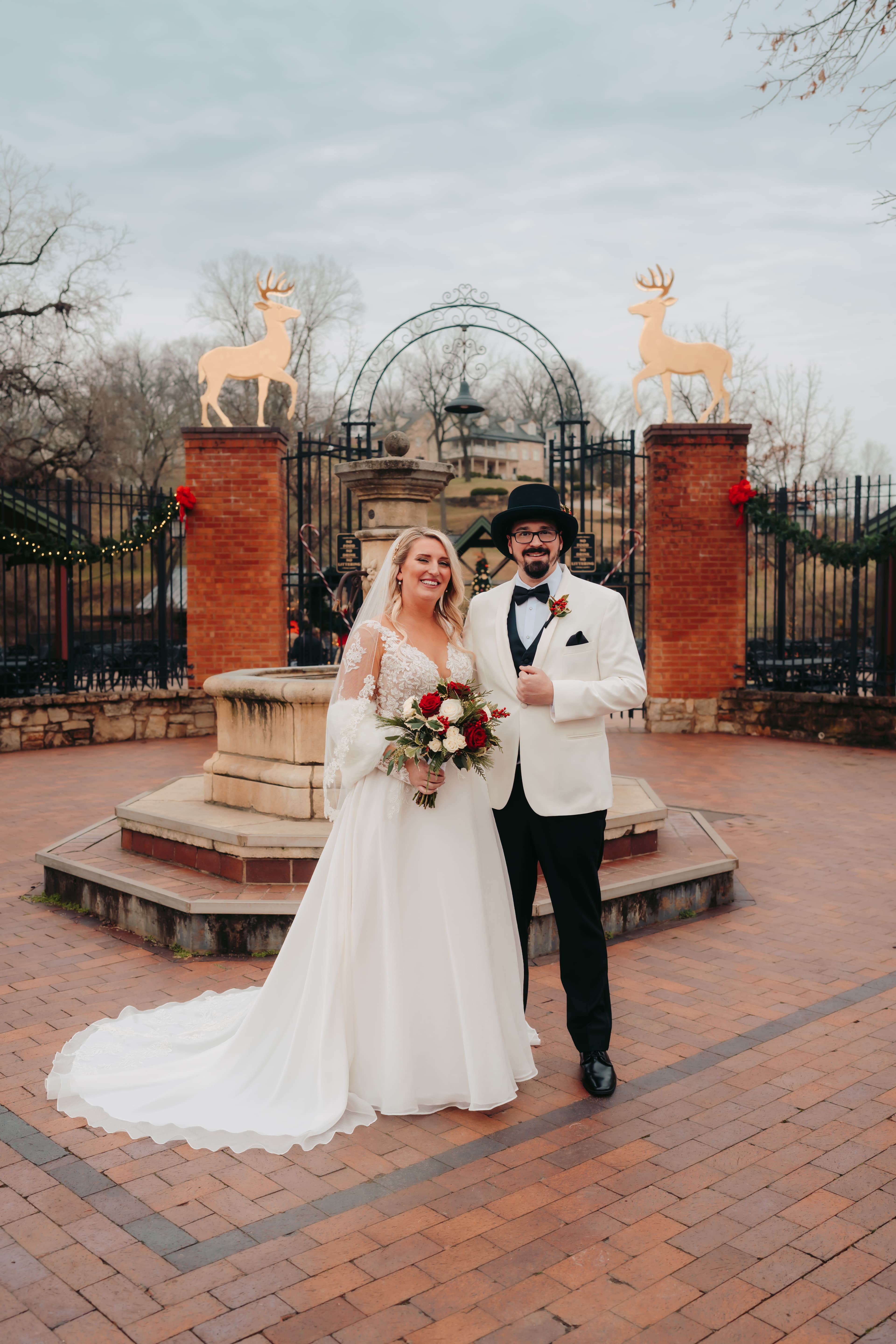 A smiling couple in formal attire stands in a scenic outdoor venue, with festive decorations and a fountain in the background.