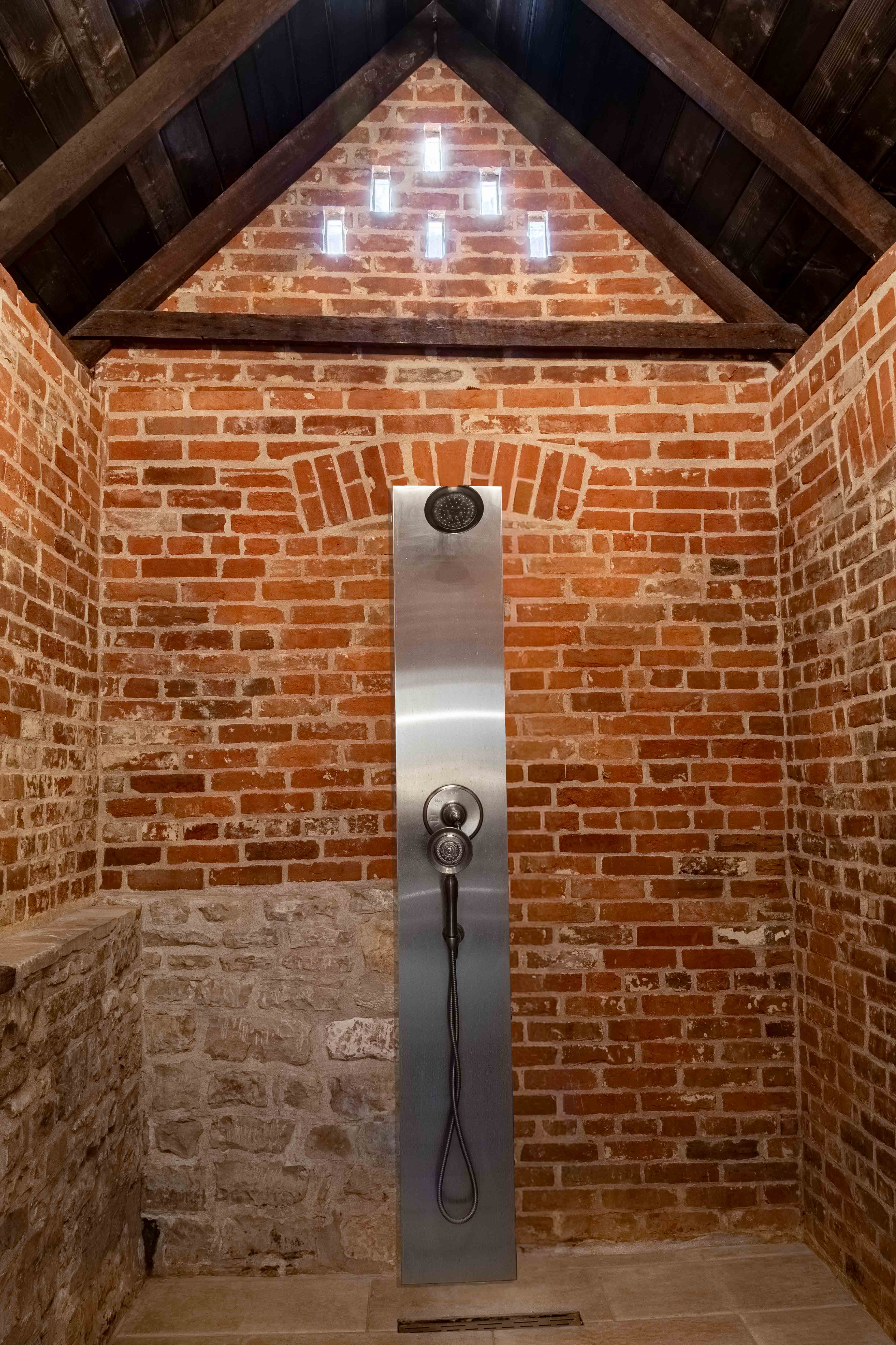 A modern shower with a stainless steel panel is set against a rustic brick wall.