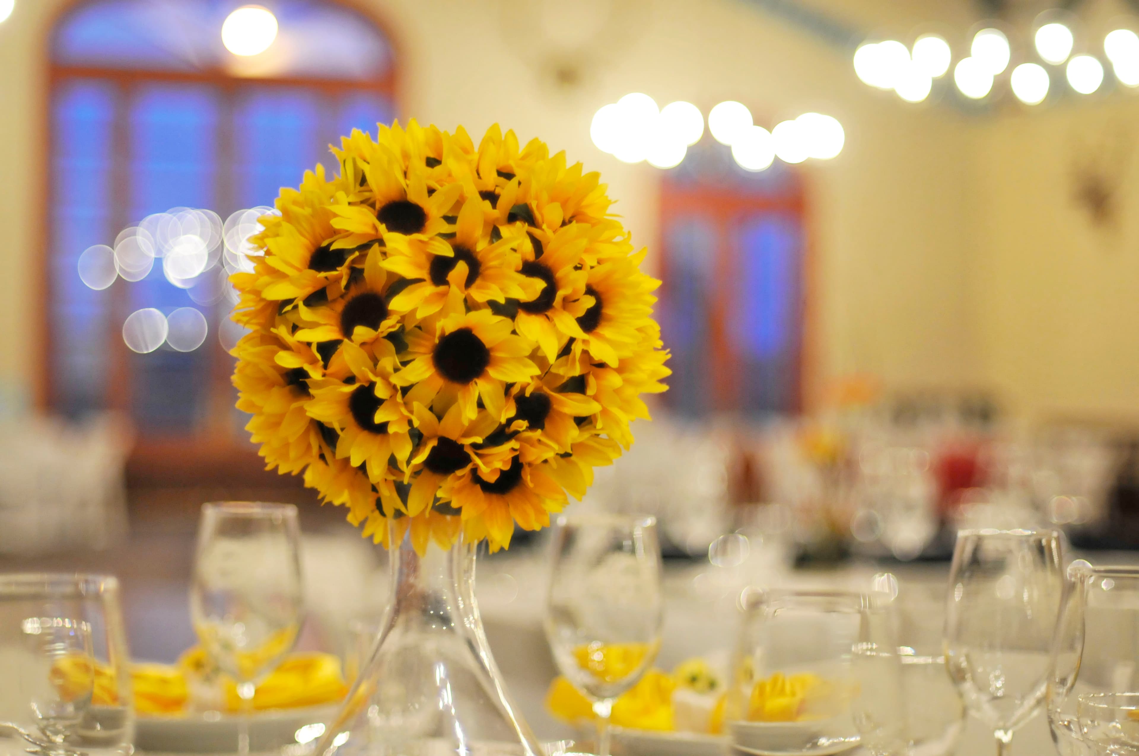 A vibrant bouquet of sunflowers in a glass vase surrounded by dining tables and softly glowing lights.