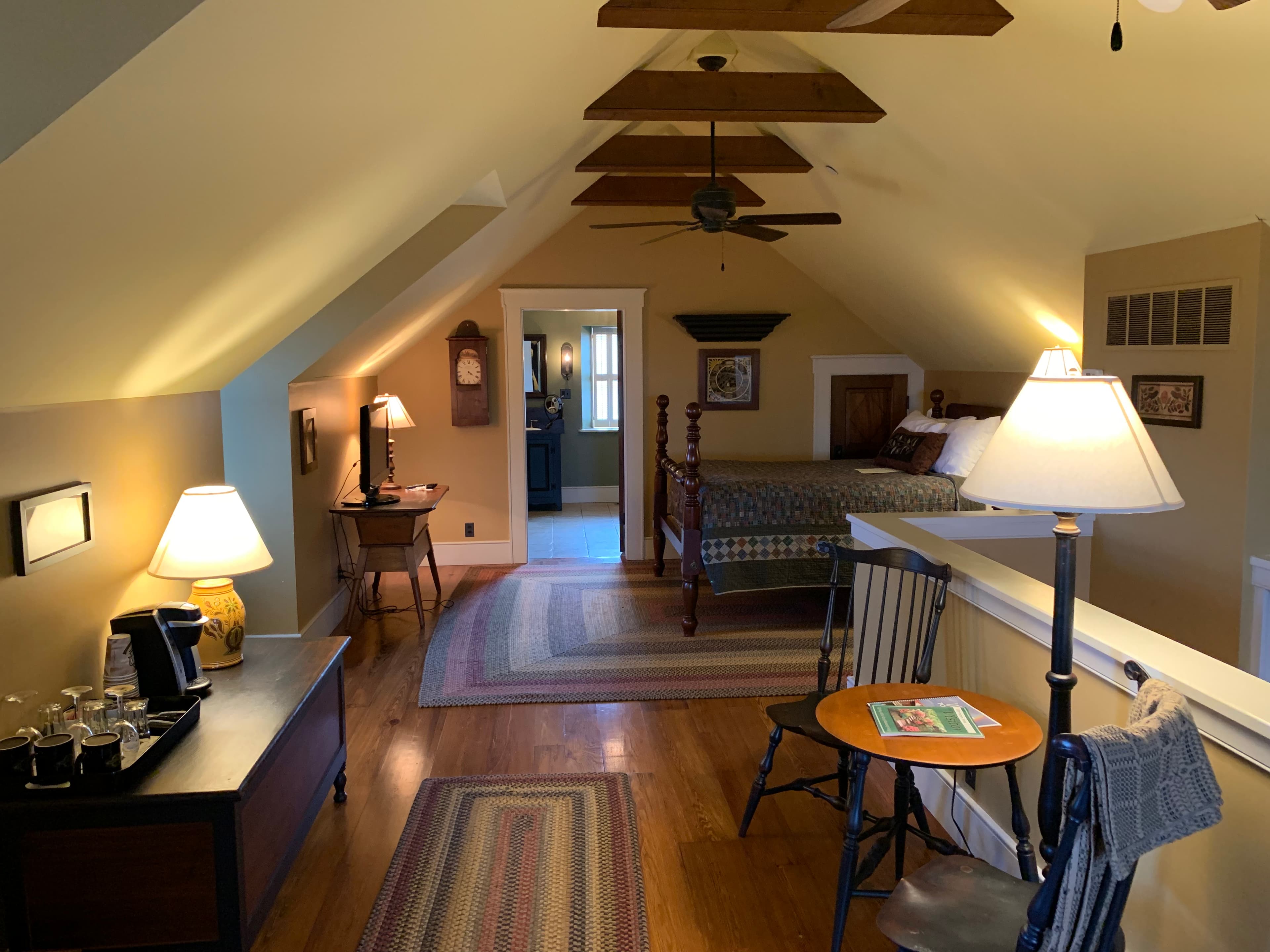Cozy attic bedroom with wooden beams, a bed, and sitting area, featuring warm lighting and rustic decor.