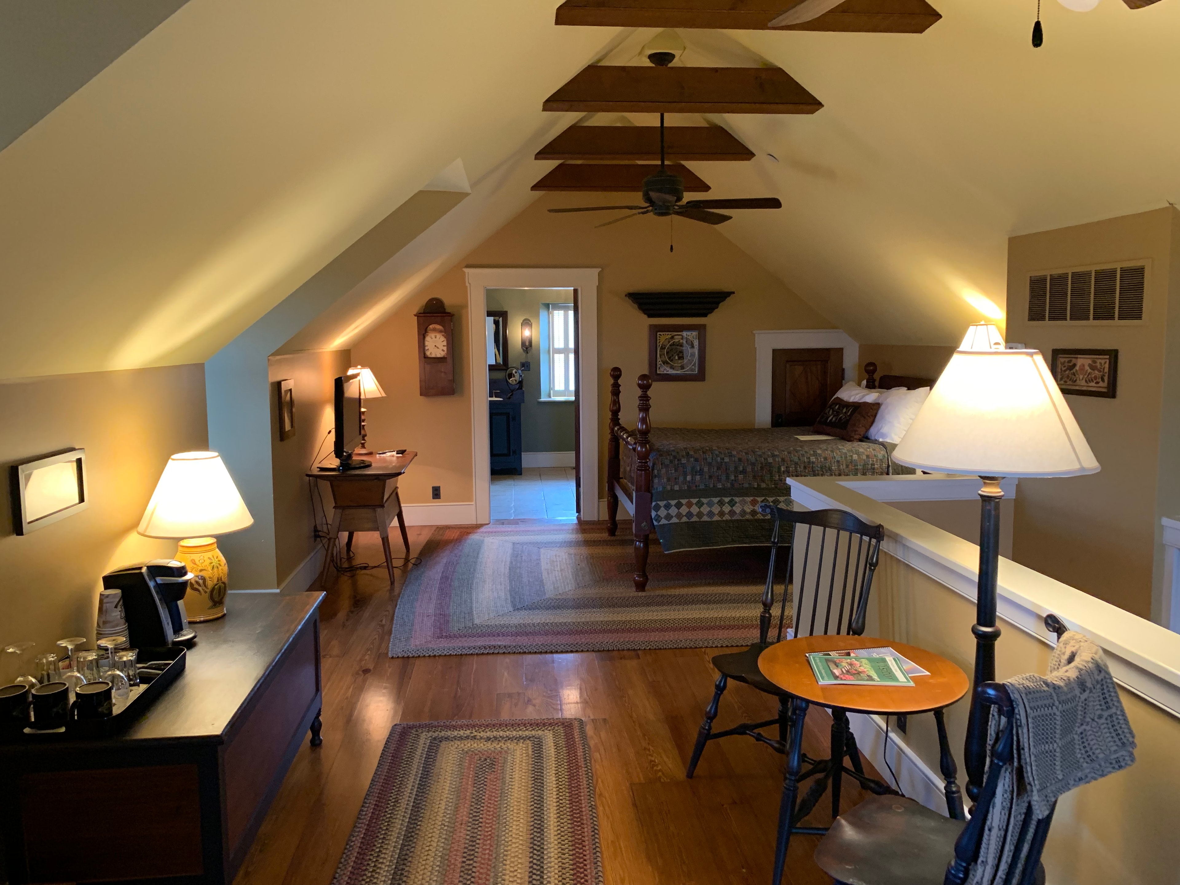Cozy attic bedroom with wooden beams, a bed, and sitting area, featuring warm lighting and rustic decor.