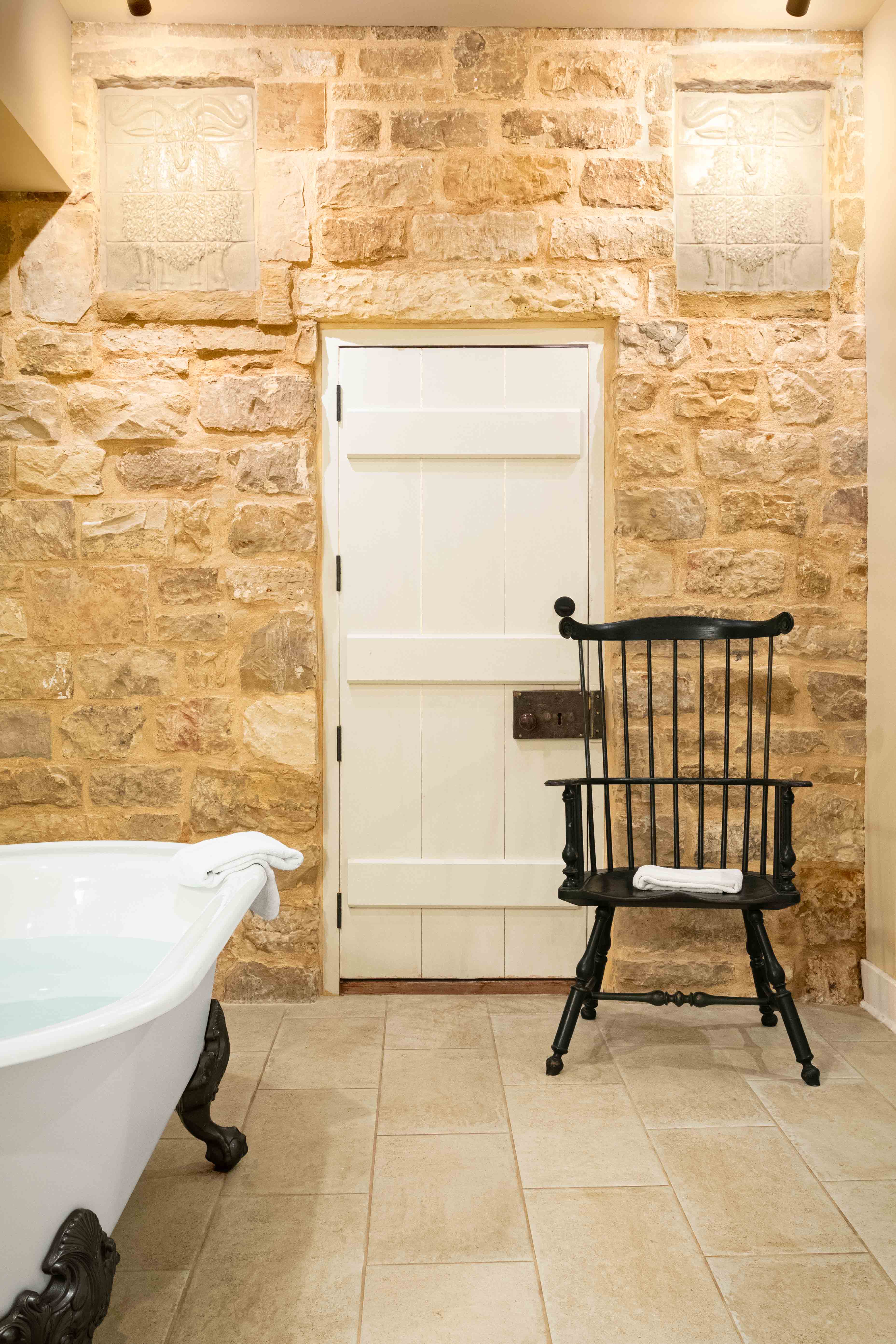 A vintage black chair stands next to a white bathtub in a stone-walled bathroom.