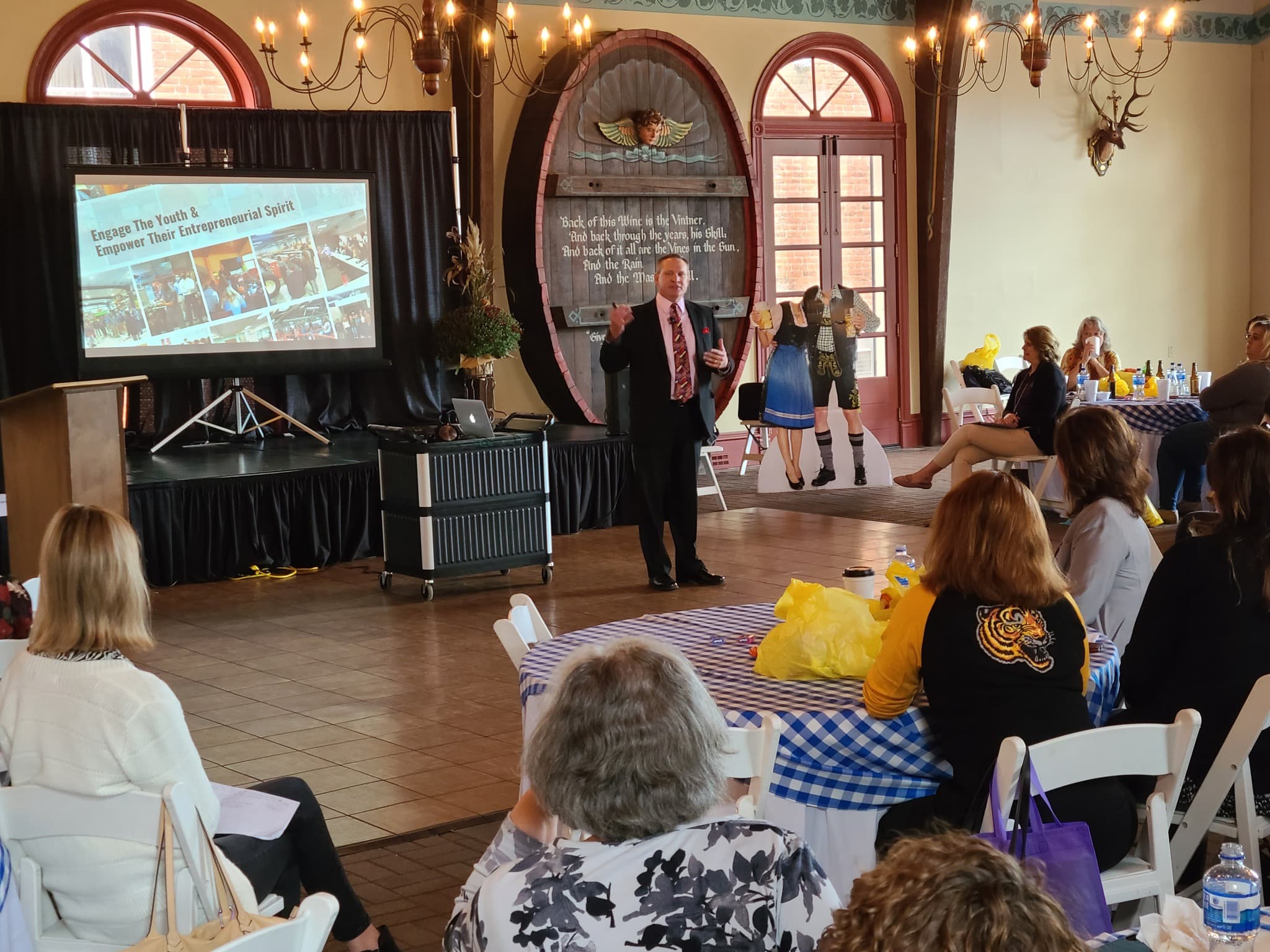 A speaker presents to an audience in a decorated venue with a large screen behind him.
