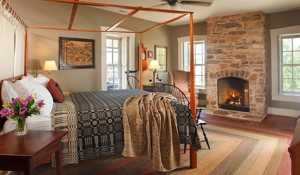 Cozy bedroom with a four-poster bed, a stone fireplace, and natural light from multiple windows.