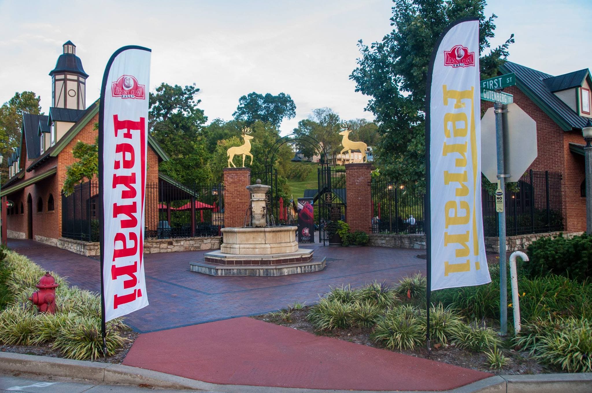 Ferrari-branded flags stand at the entrance of a plaza featuring a fountain and landscaped greenery.