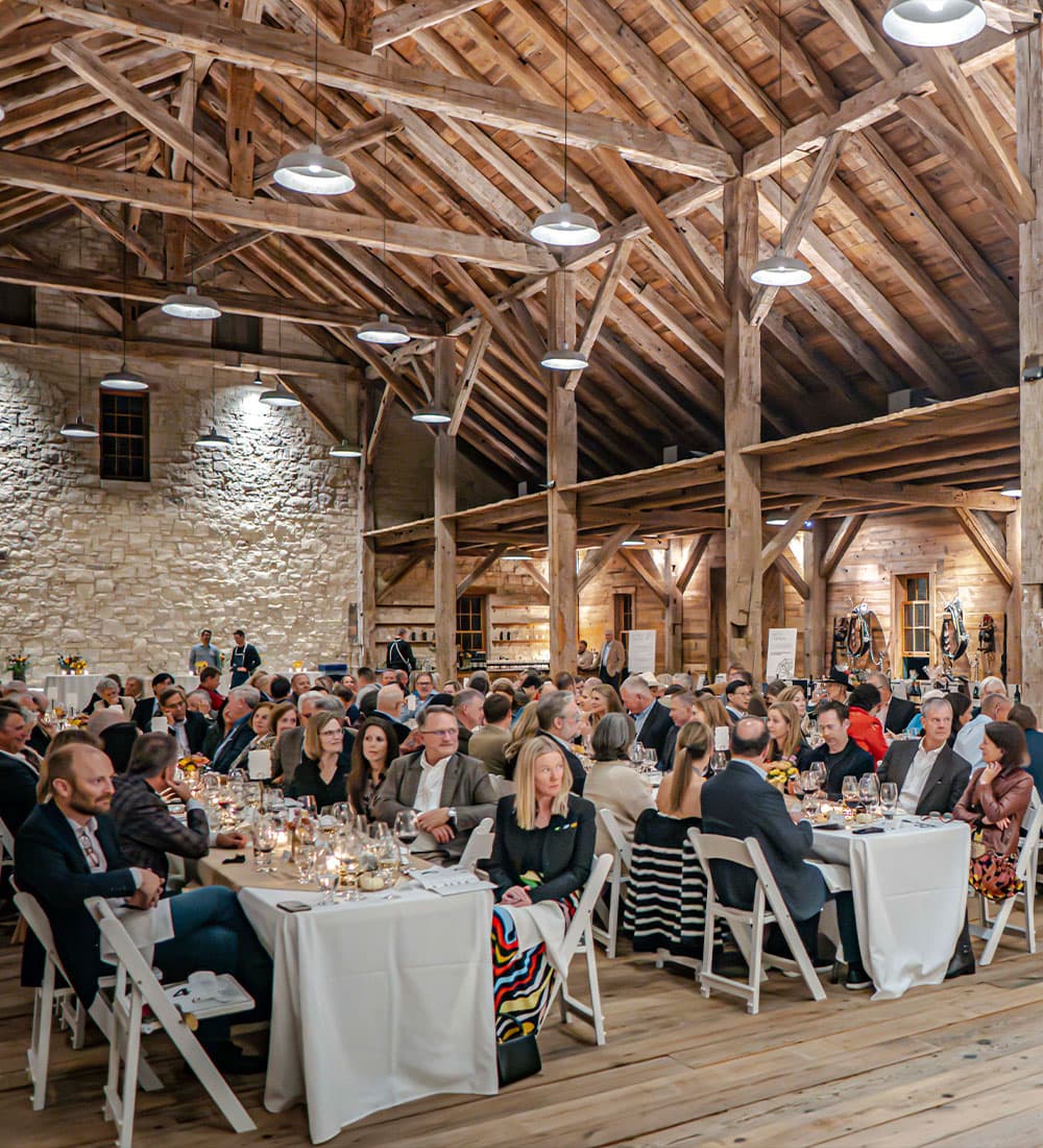 A large group of elegantly dressed guests sits at tables in a rustic barn setting, enjoying a banquet.