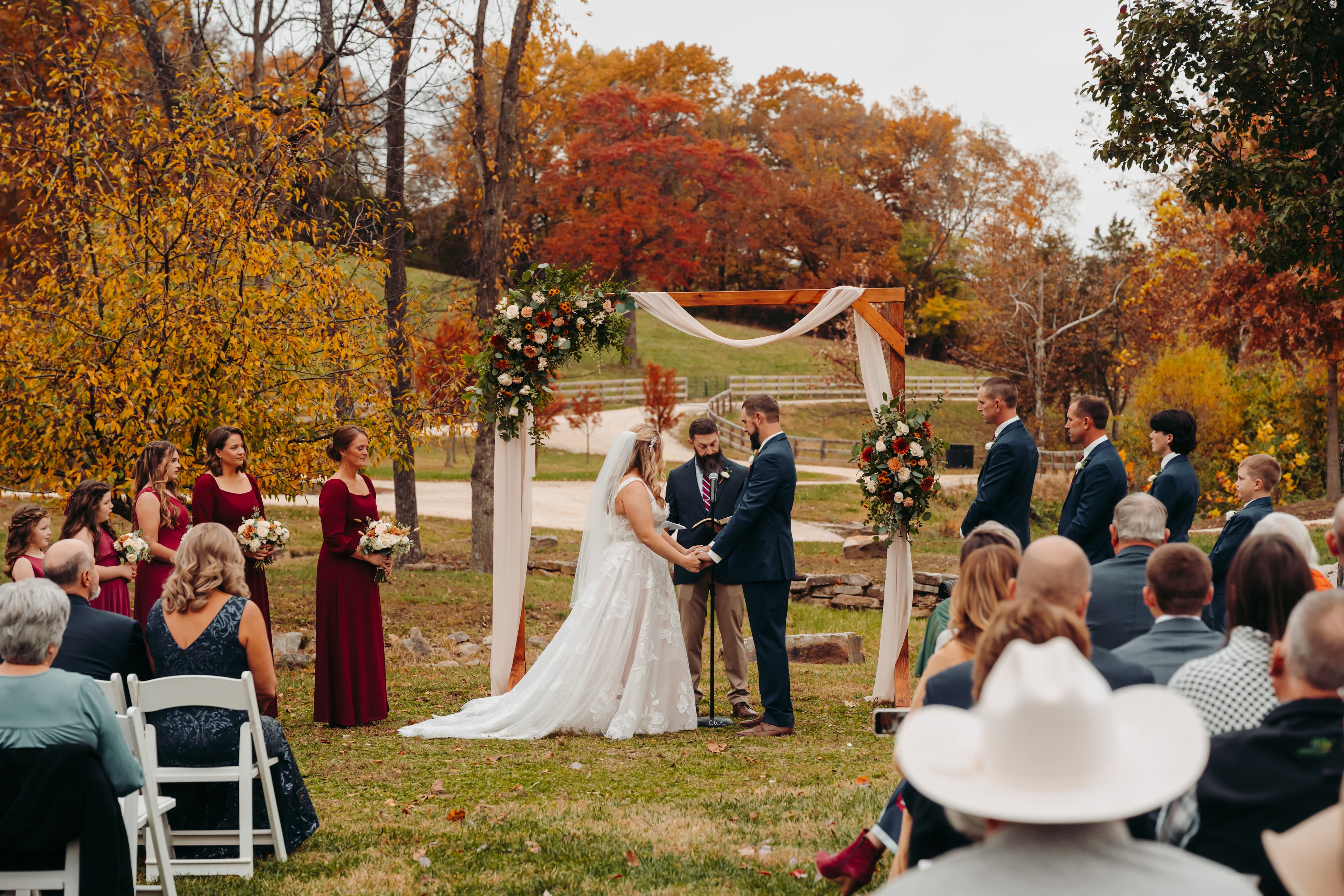 A couple exchanges vows at an autumn outdoor wedding, surrounded by guests and floral decorations.