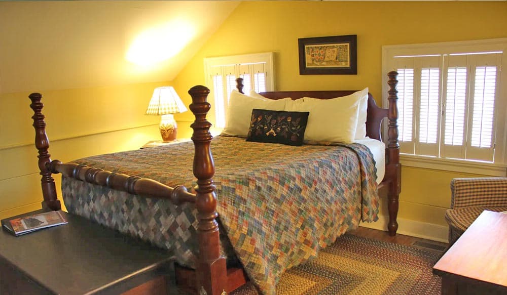A cozy attic bedroom featuring a wooden bed, colorful quilt, bedside lamp, and a patterned chair.